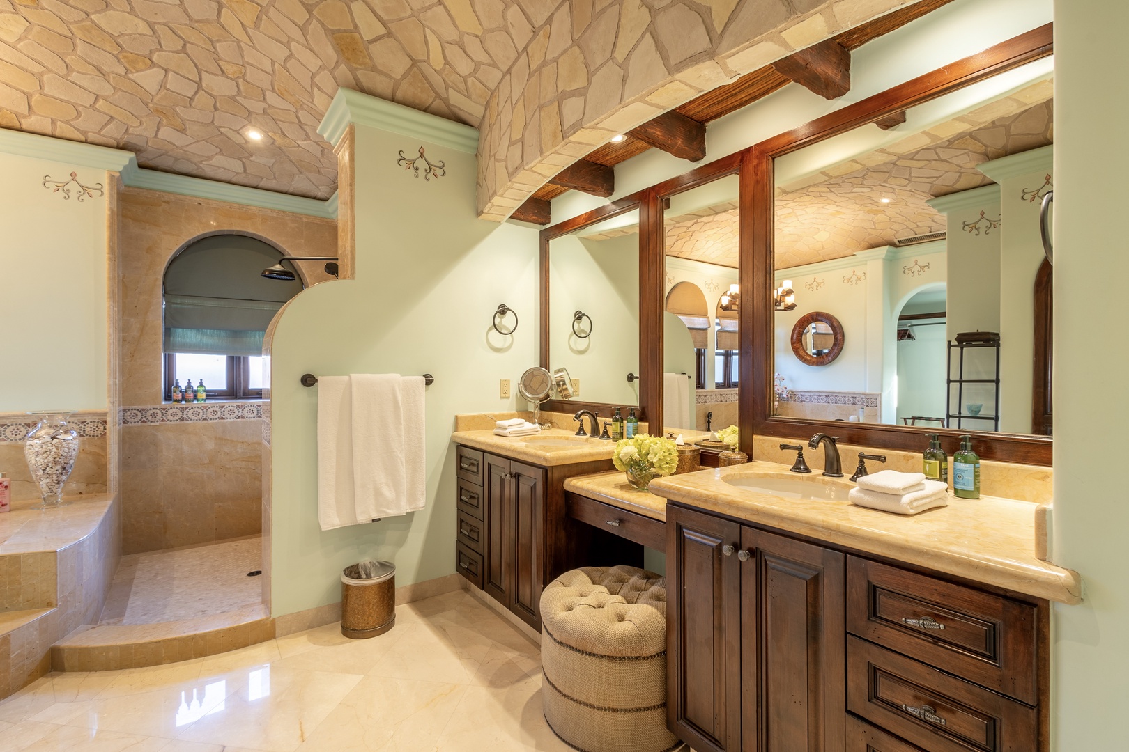 Unwind in your elegant bathroom retreat featuring rustic wood beam ceilings, stone archways, dual vanities, and plush towels awaiting your refreshing shower.