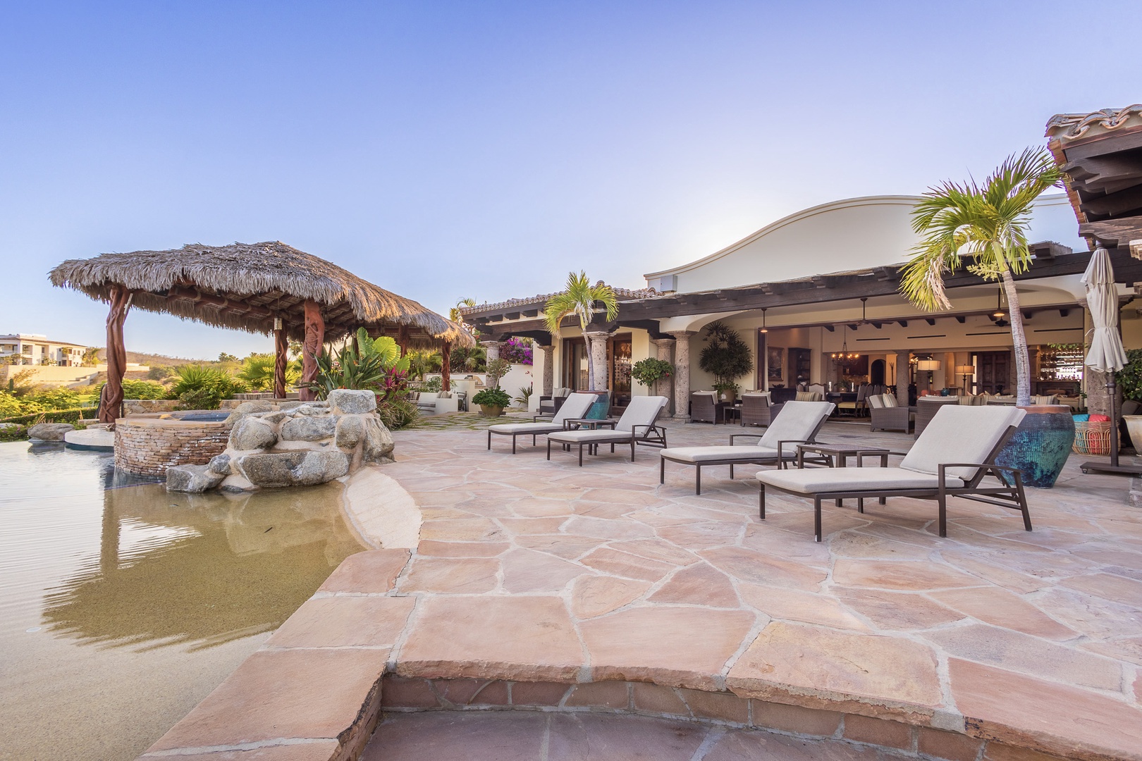Private beachfront villa featuring tropical palapa shade, decorative pond, and poolside loungers surrounded by lush palms and desert hillsides.
