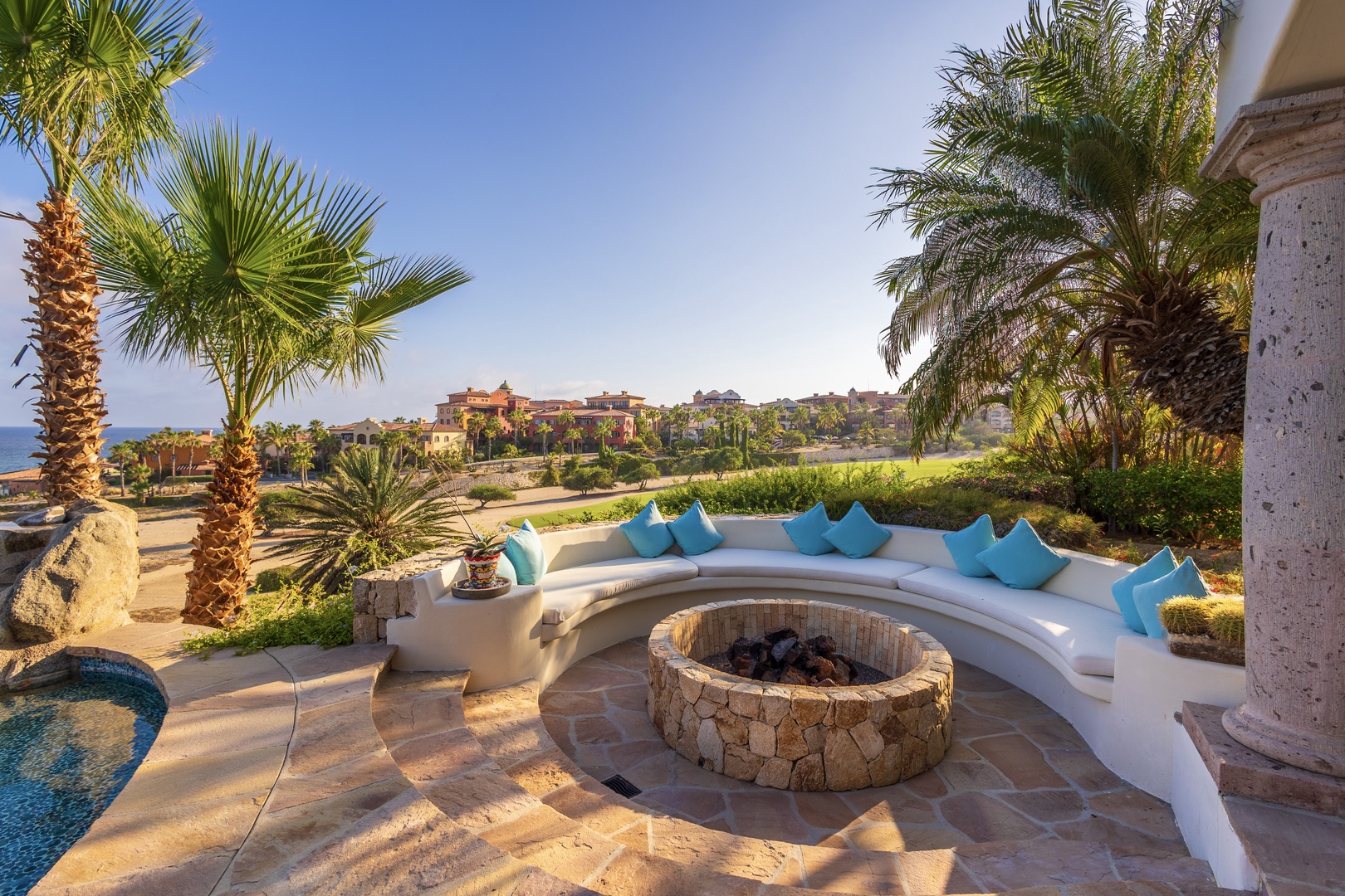 Step outside to your private oasis where coastal breezes meet luxurious comfort. Lounge on curved seating with vibrant pillows, gather around the stone fire pit, and soak in sweeping ocean and garden views framed by swaying palms.