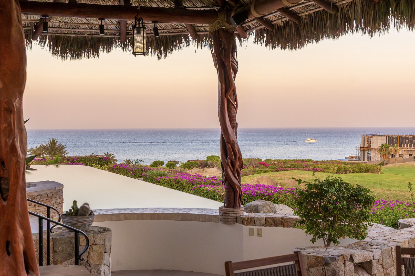 Seaside terrace framed by tropical palapa offers stunning sea views with vibrant gardens, green lawn, and sunset skies—an idyllic spot to unwind and soak in coastal beauty.