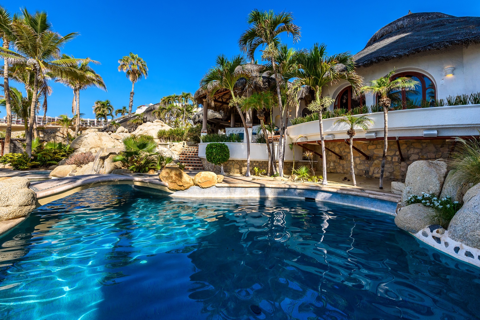 Stunning resort-style pool framed by swaying palms, natural rock formations, and thatched-roof architecture under clear blue skies—a private oasis for total relaxation.