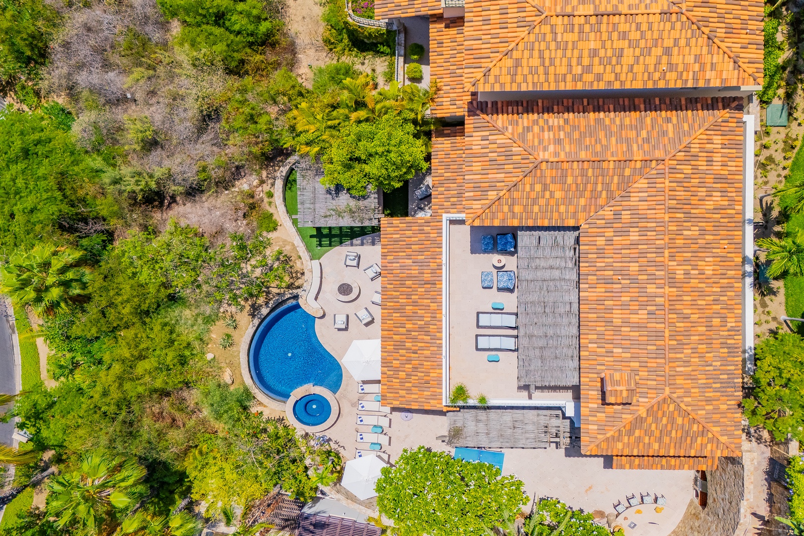Aerial view of a stunning vacation property featuring a distinctive terracotta-tiled roof, curved swimming pool with adjacent spa, and surrounding lush natural landscape.