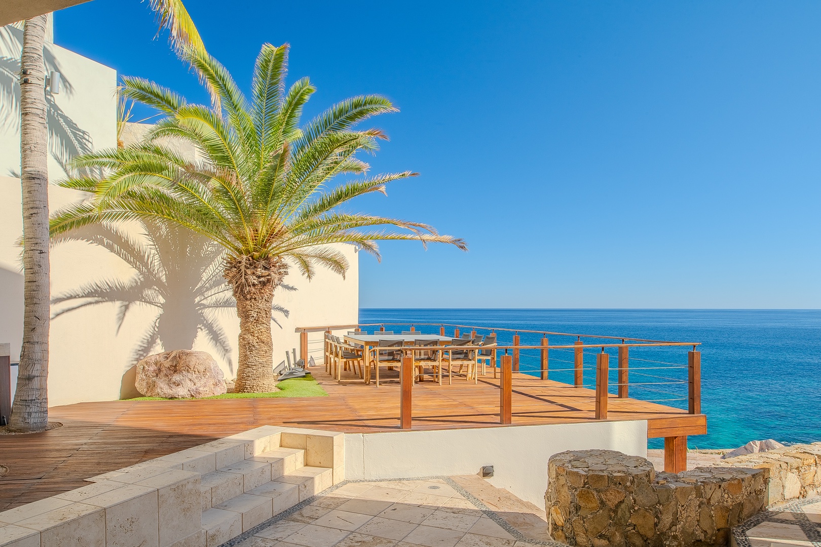 Seaside wooden deck with dining space, palm tree shade, and direct views of turquoise waters under clear skies—perfect for coastal relaxation.