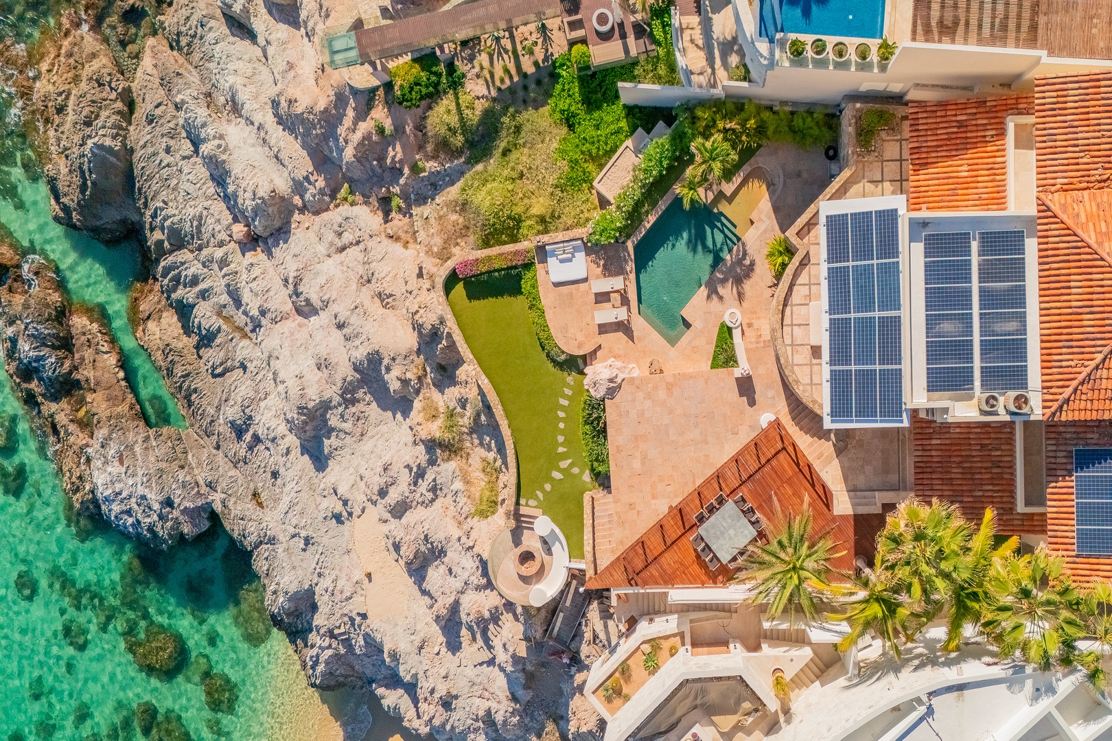 This coastal retreat perches dramatically on rugged cliffs above turquoise waters. The property features terracotta roofs, manicured lawns, a private pool, and palm-fringed terraces overlooking the stunning seascape.