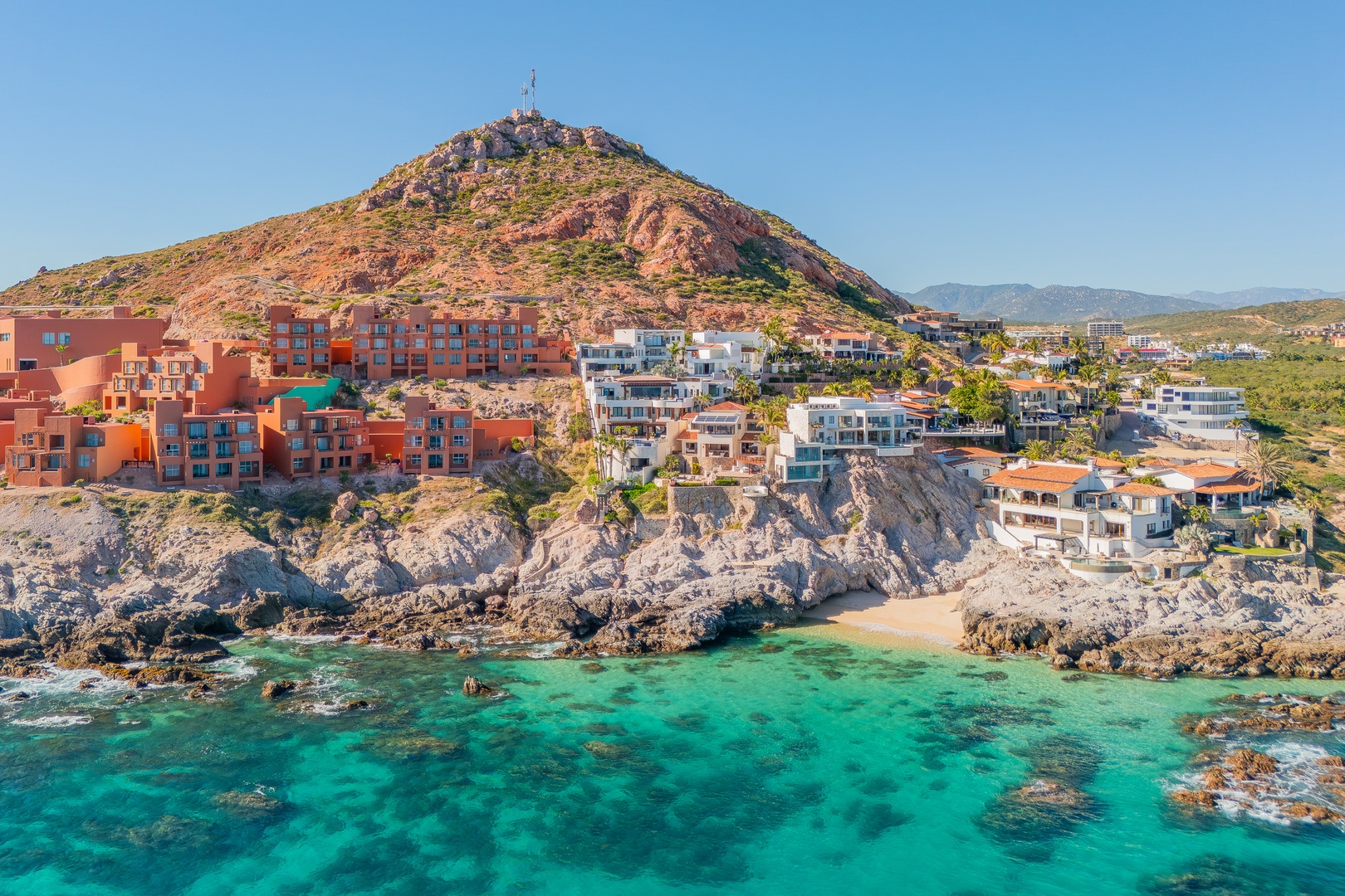 Coastal resort community nestled against rocky hillside with turquoise waters, sandy beach, and colorful buildings cascading down to the shore under clear blue skies.