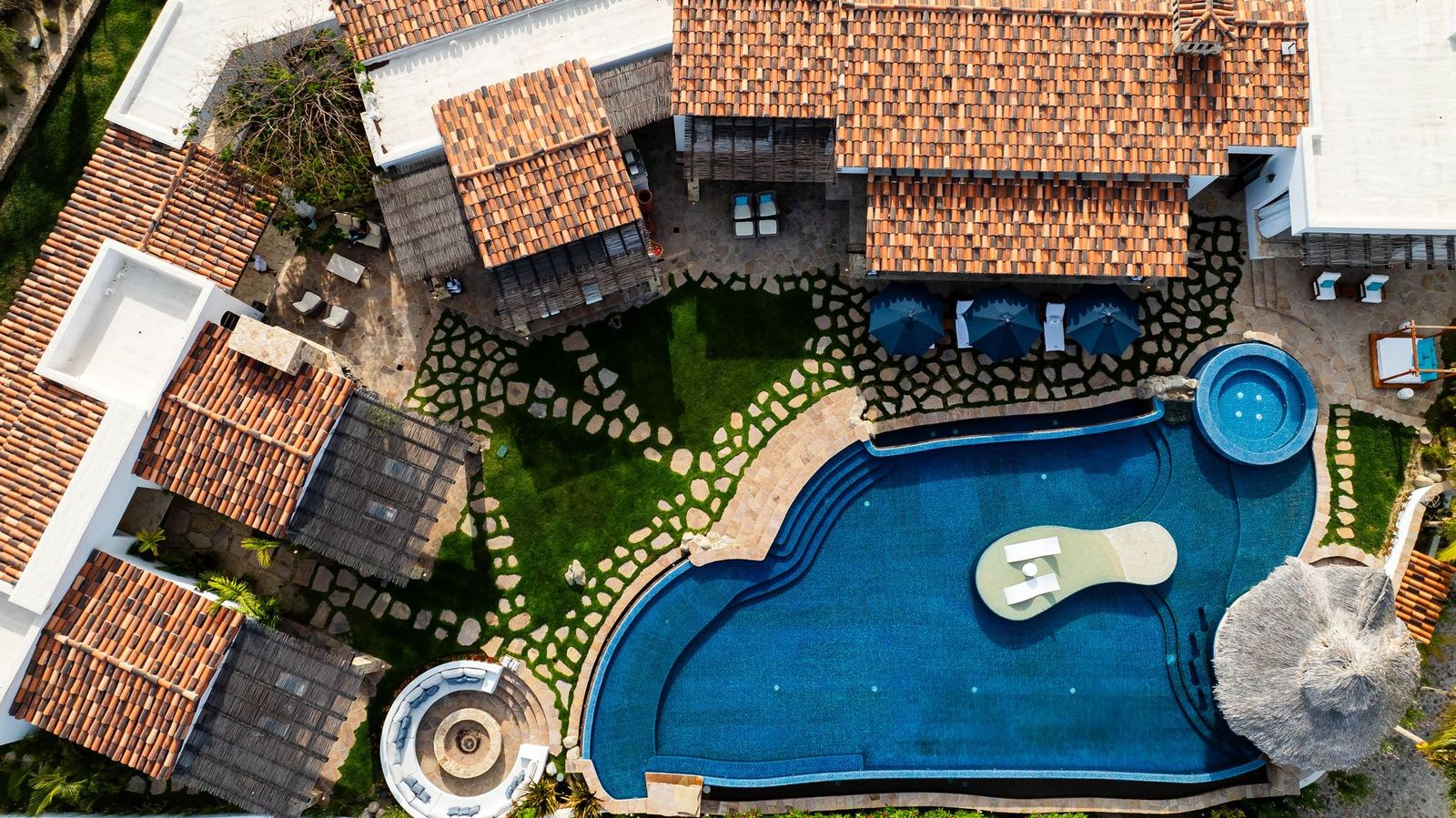 Aerial view showcasing the property's expansive pool area with integrated hot tubs, sunbeds, and multi-level terracotta-roofed buildings set within beautifully landscaped grounds.