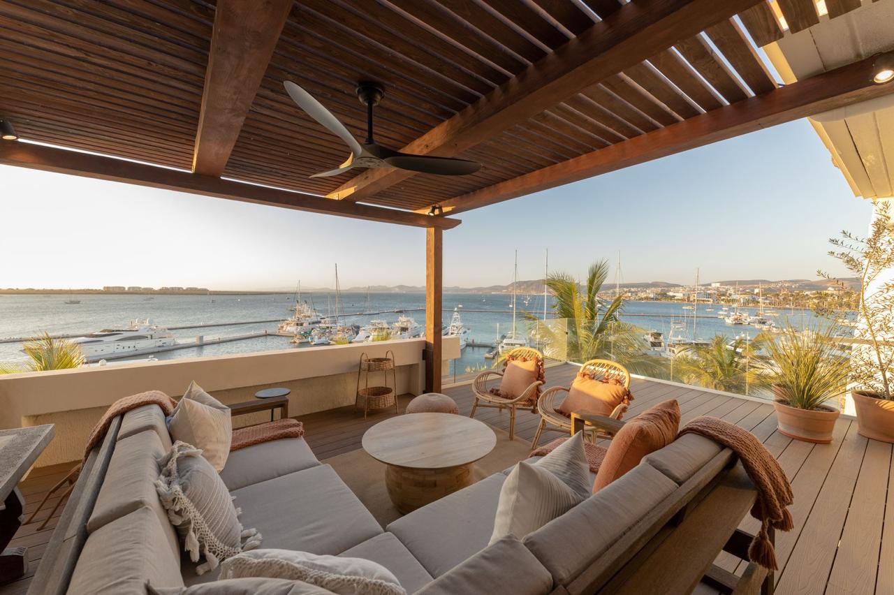 A sophisticated covered terrace overlooking a marina and coastal vista, featuring plush seating, warm wood details, and a ceiling fan for comfort. Ideal for relaxing while watching boats drift by.