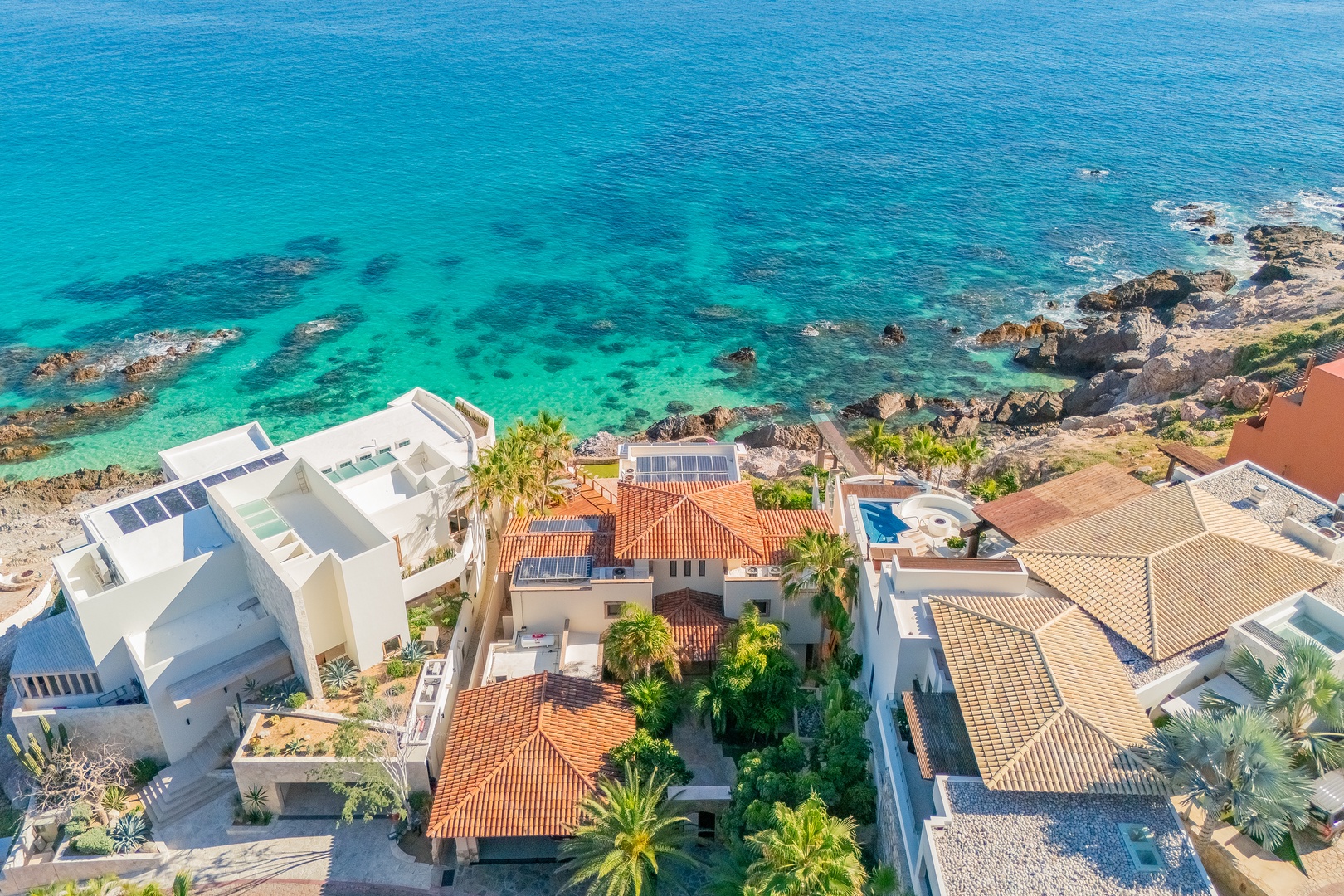 Aerial view showcasing coastal villas with terracotta and contemporary rooflines nestled along a stunning turquoise shoreline with rocky outcrops and crystal-clear waters.