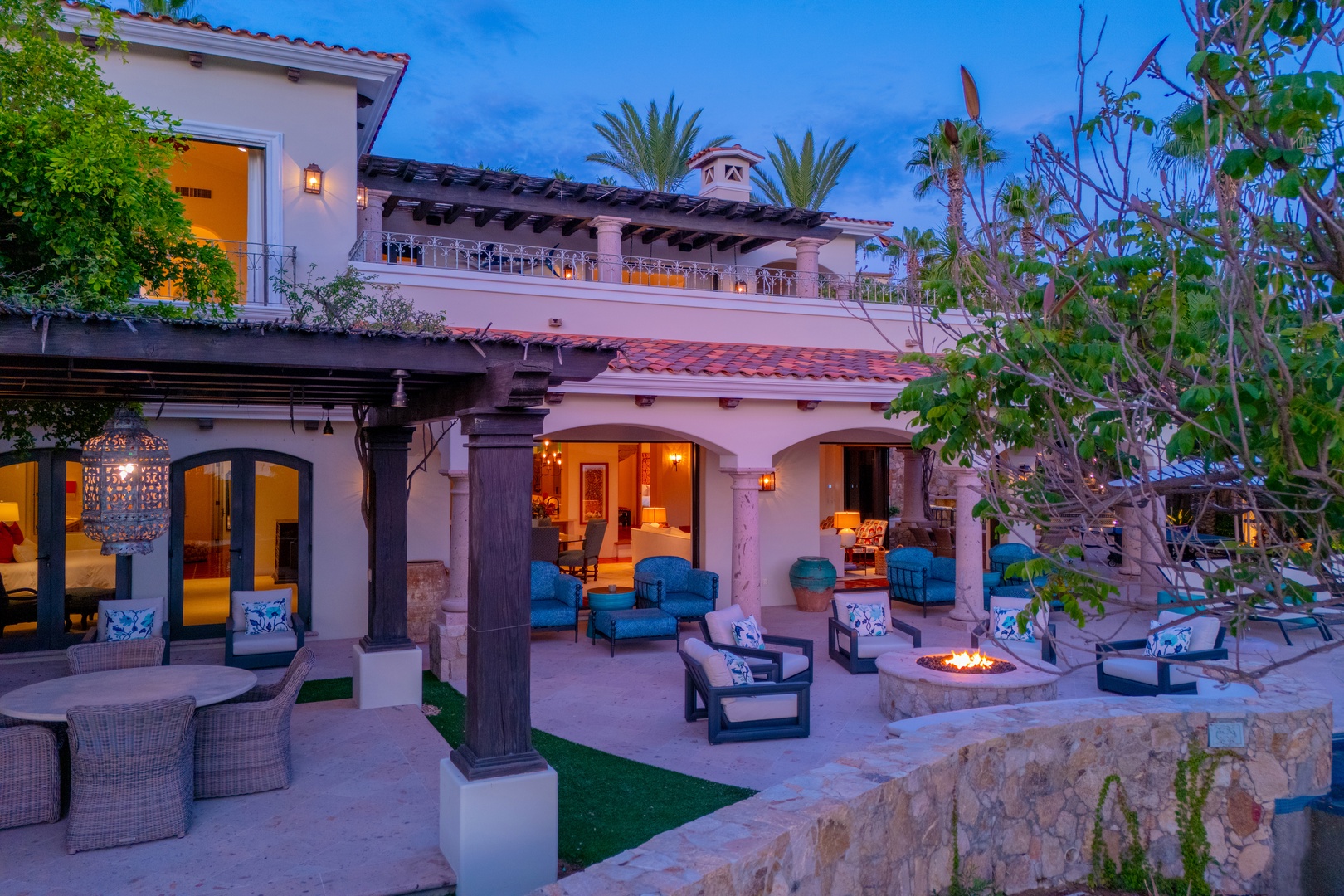 A stunning Mediterranean-style villa photographed at twilight, featuring multi-level architecture with red-tile roofing, arched doorways, and lush tropical landscaping. The property's elegant outdoor living spaces are softly illuminated against the evenin