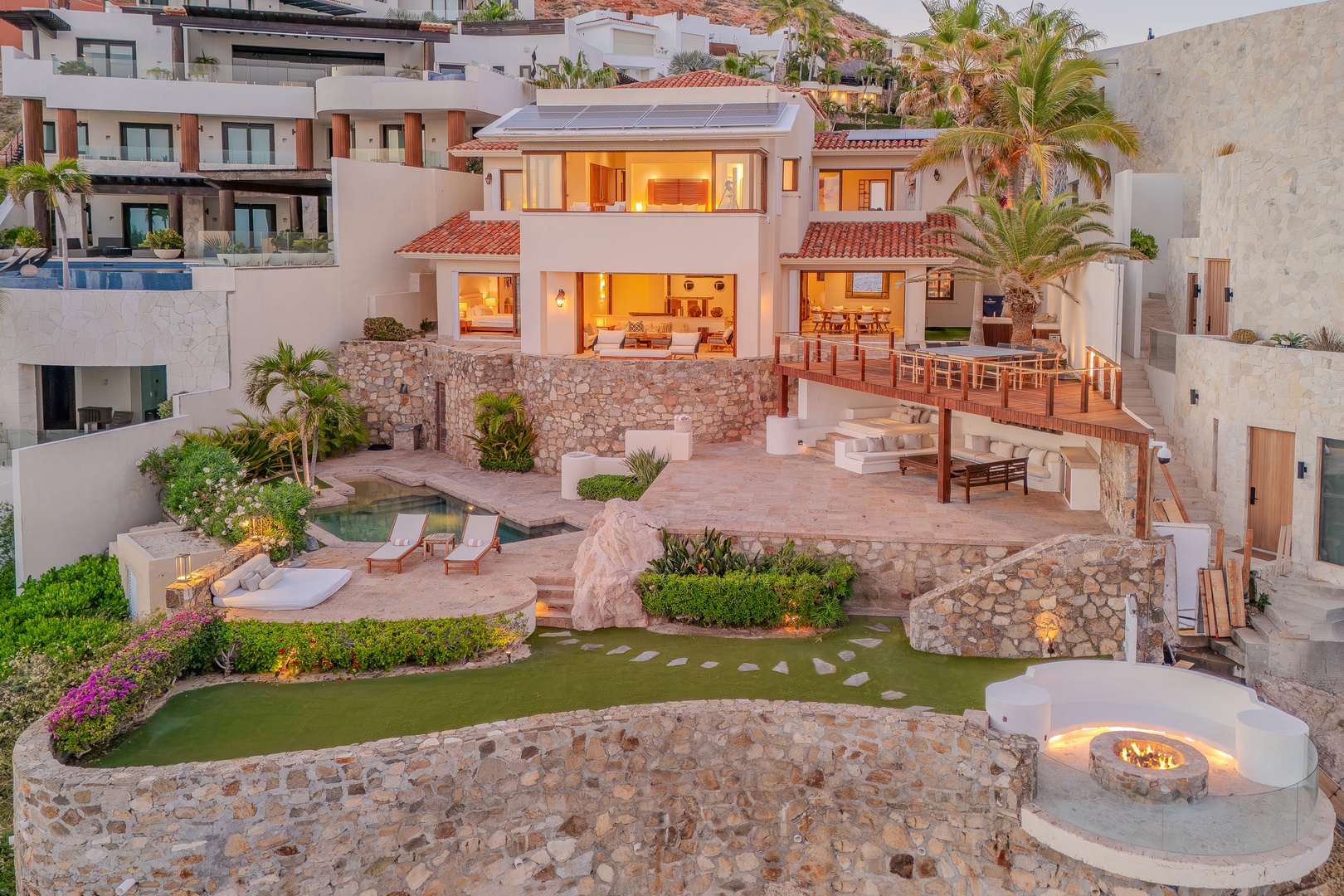A stunning multi-level villa showcases illuminated outdoor living spaces with terraced patios, private pool, fire pit, and lush tropical landscaping at dusk.