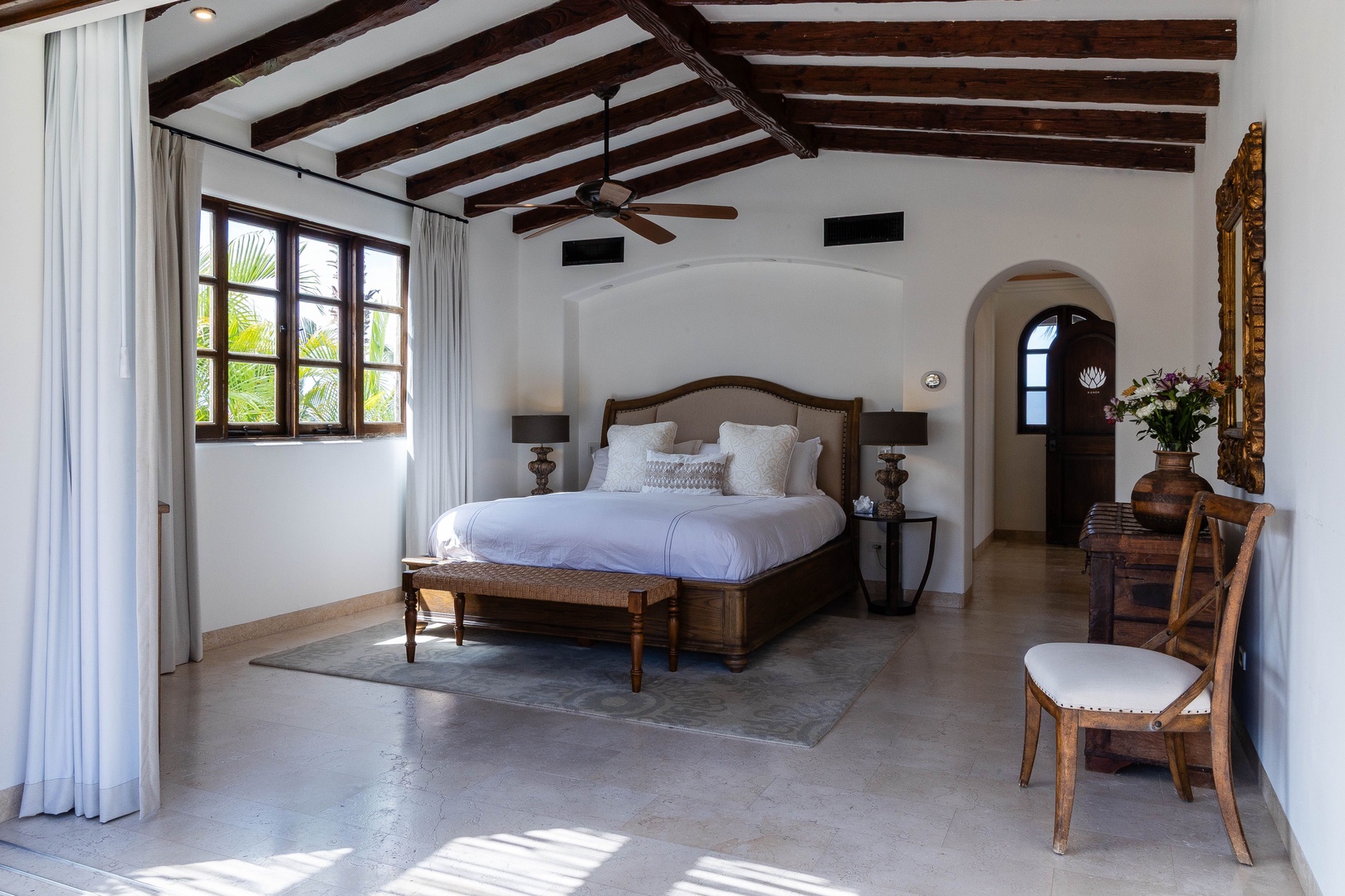 Your spacious bedroom blends rustic charm with elegant comfort, featuring exposed beams, a plush bed, and arched doorways that open to peaceful garden views.