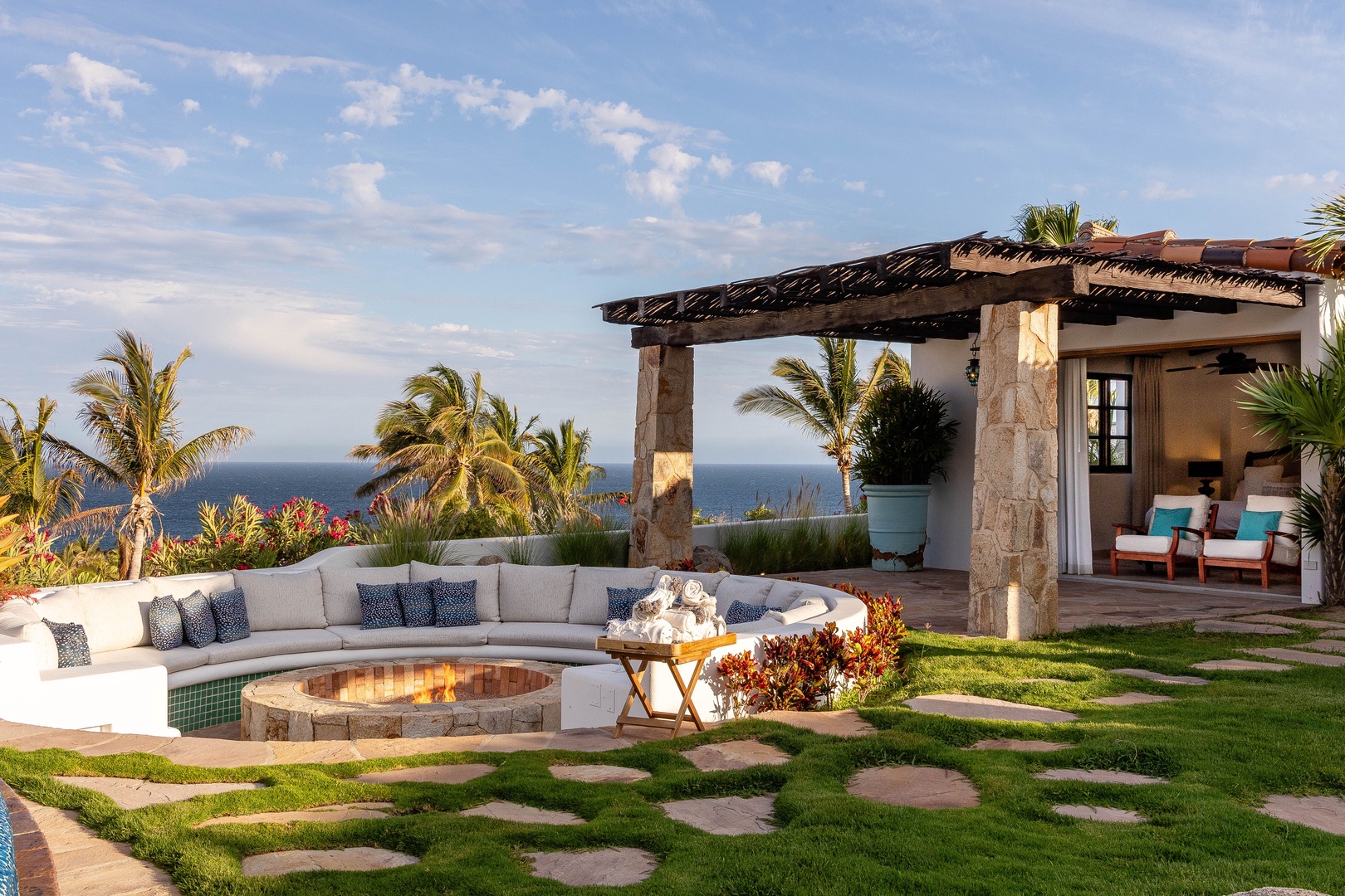 This oceanfront retreat features a circular stone fire pit surrounded by curved seating, stone pathways through manicured lawns, and a covered outdoor lounge overlooking swaying palms and endless sea views.