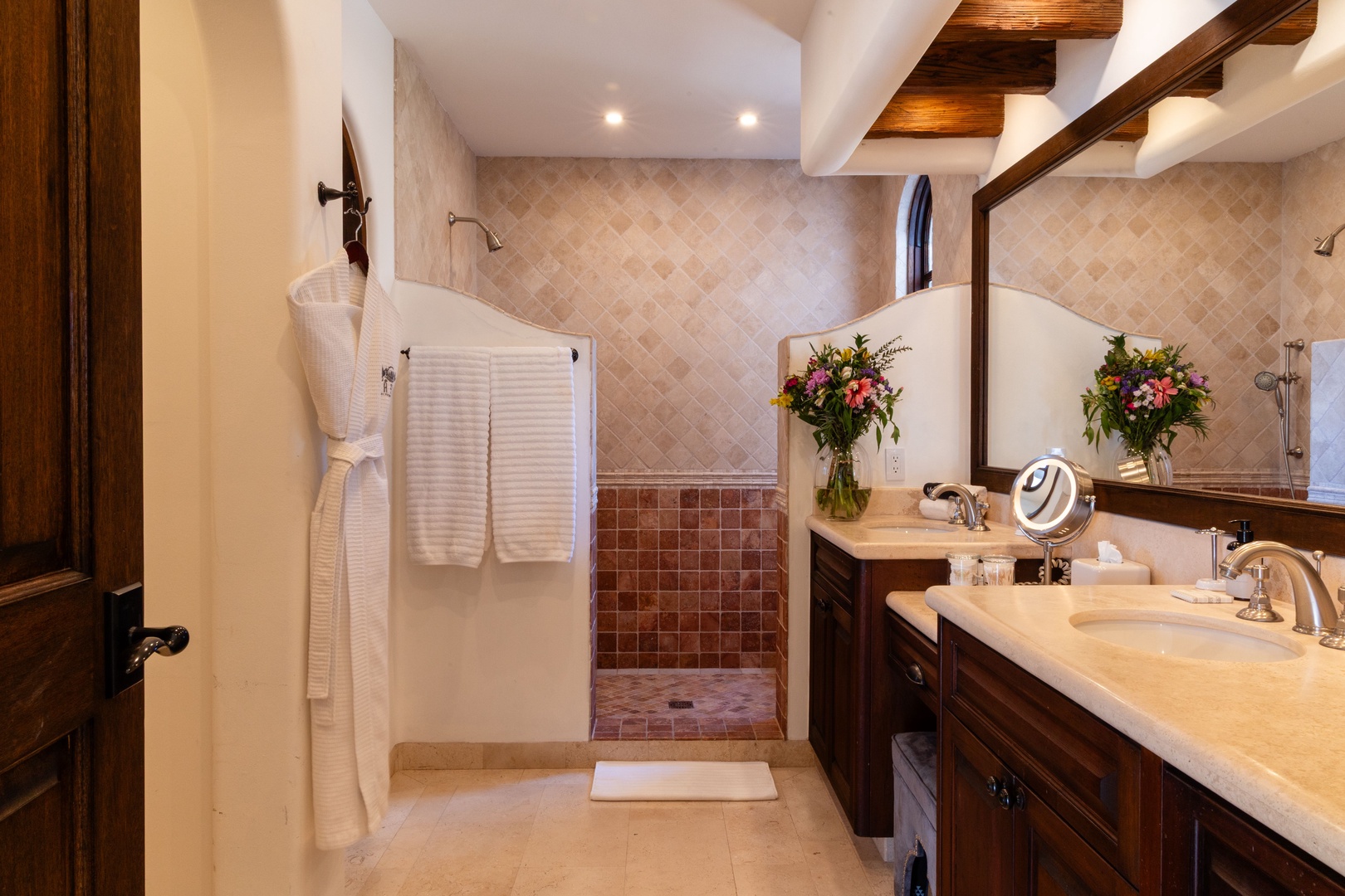 Refresh in your private bathroom with double sinks, fresh flowers, and a spacious walk-in shower—rustic wood beams add warmth overhead.
