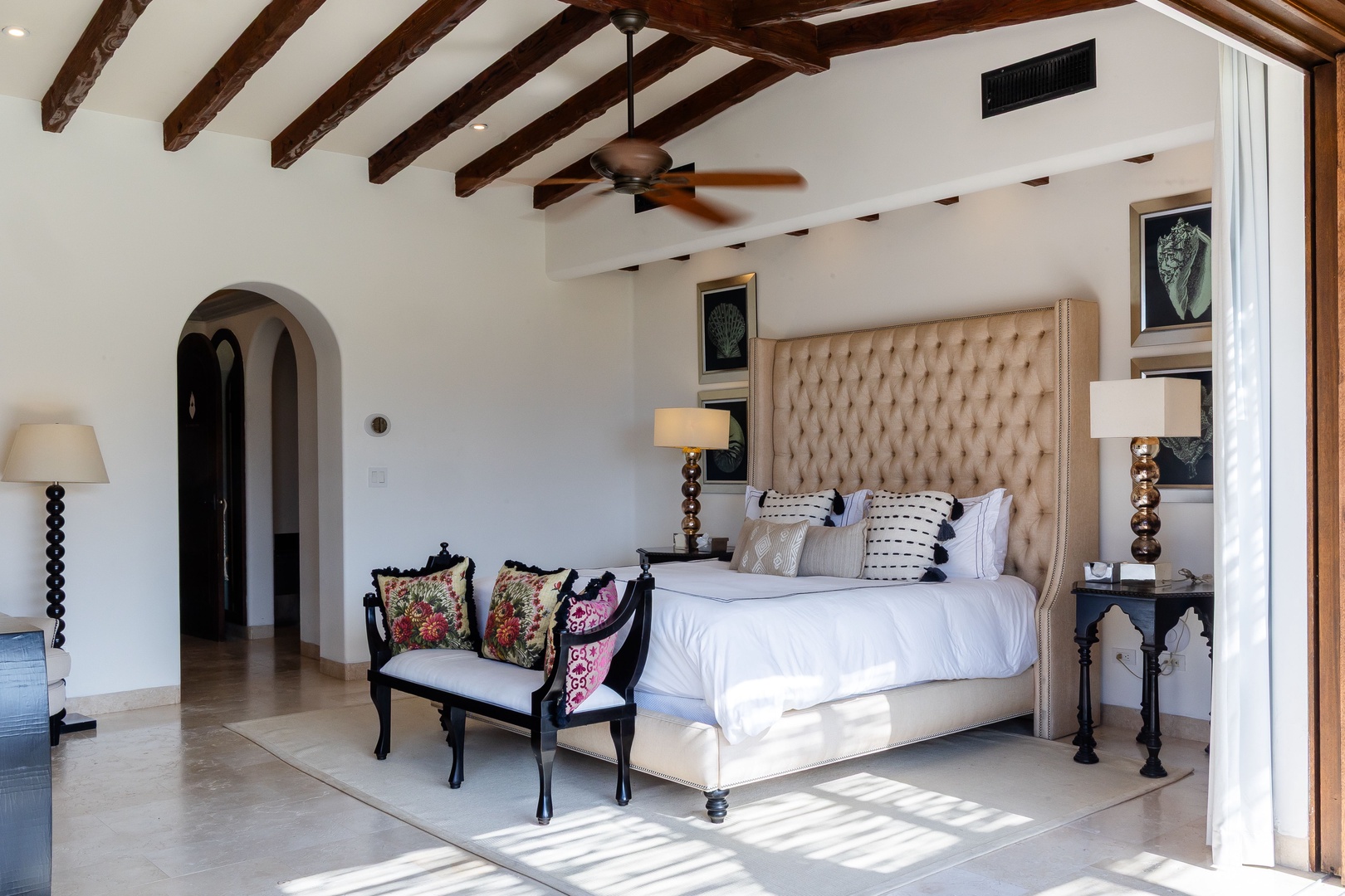 Step into your elegant bedroom, where exposed wood beams frame soaring ceilings and natural light dances across gleaming tile floors. Sink into plush bedding beneath a statement tufted headboard, with stylish seating and warm lighting creating your perfec