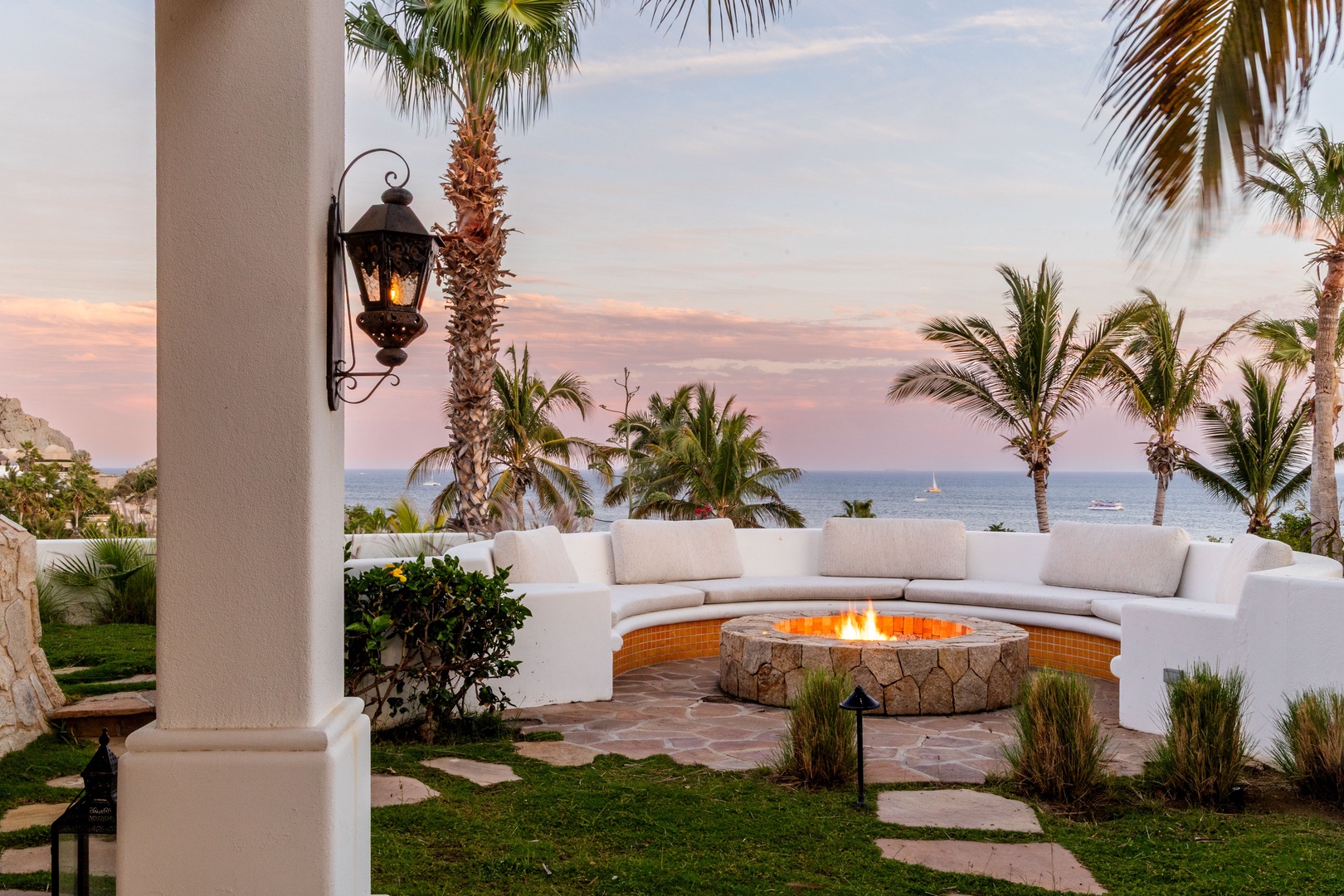 Coastal retreat featuring elegant outdoor seating around a fire pit, framed by palm trees and ocean views at sunset.