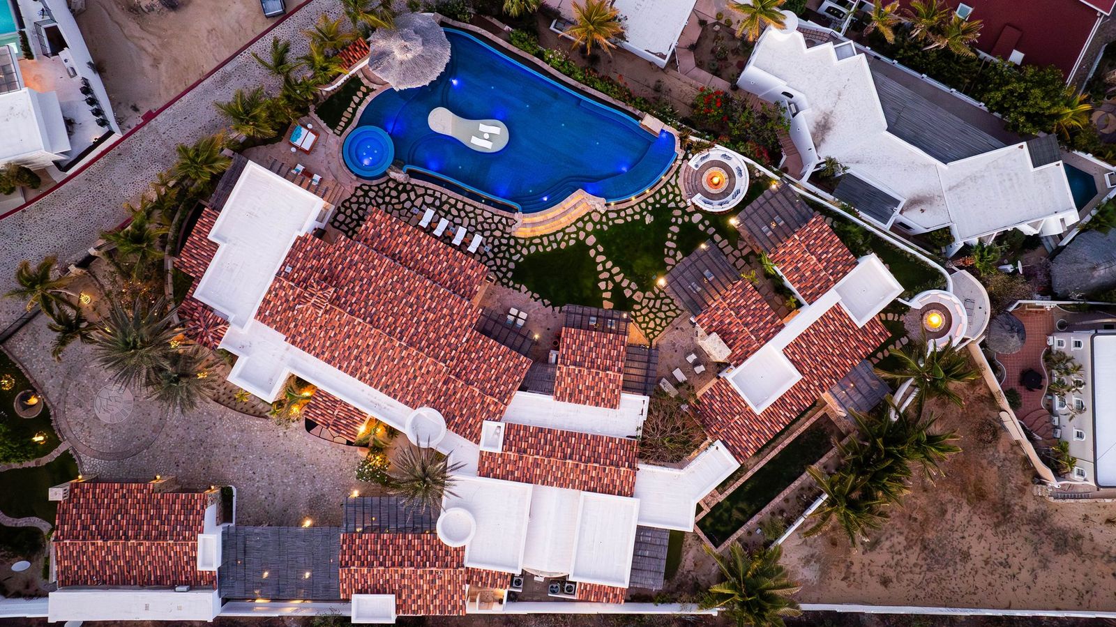 Aerial view showcasing the property's distinctive architecture, terracotta roofing, blue-lit swimming pool, and lush palm-studded grounds nestled within the surrounding neighborhood.
