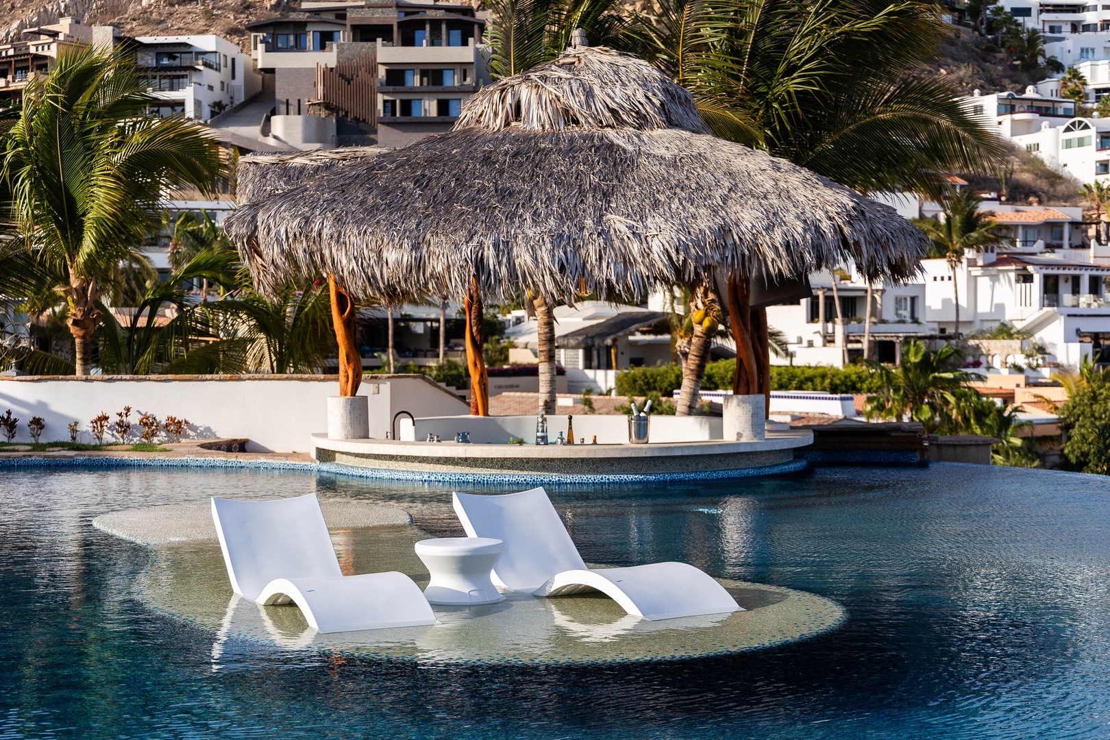 A resort-style pool features an island swim-up bar with thatched palapa and in-water loungers, set against hillside homes and palm trees.