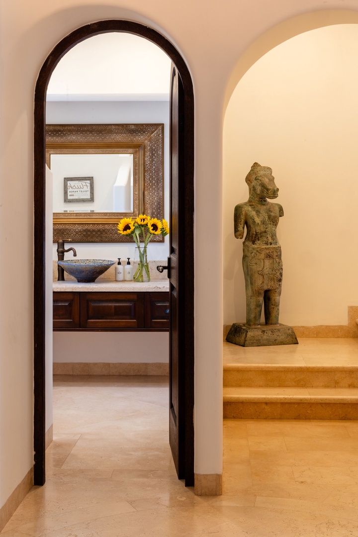 Elegant archway leads to a serene powder room with vessel sink and fresh sunflowers, while a striking stone sculpture adds cultural charm to your private retreat.