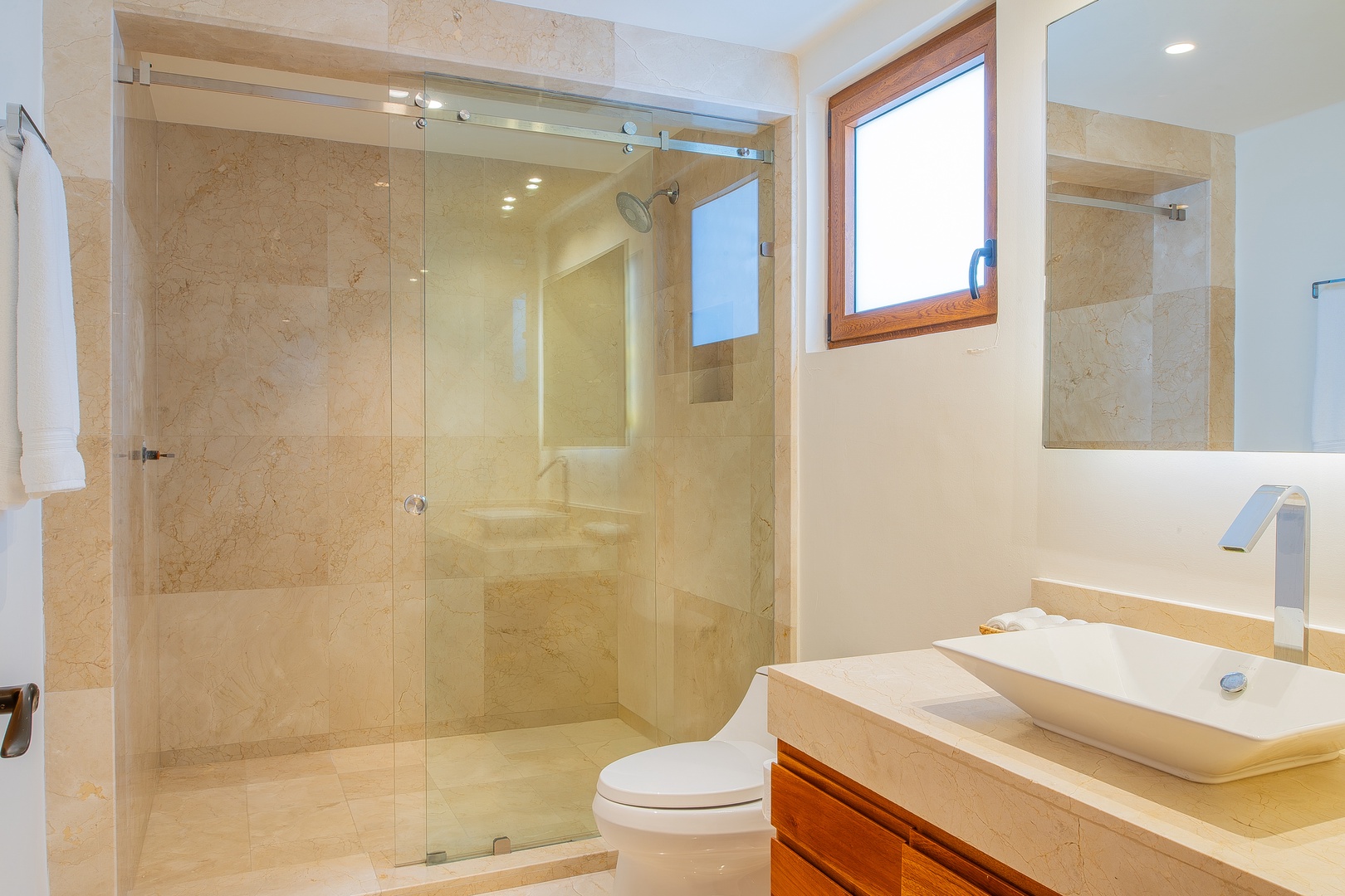 Step into your spa-like bathroom featuring warm travertine tile, a sleek modern vanity, and a spacious glass-enclosed shower. Natural light pours in through the wood-framed window, setting the perfect mood.