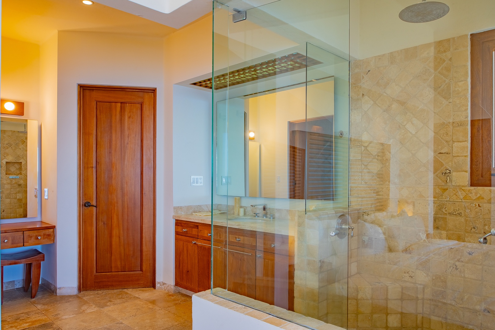 Your private bathroom features elegant travertine tile, a spacious glass-enclosed shower with rainfall showerhead, and warm wood cabinetry for a spa-like retreat.