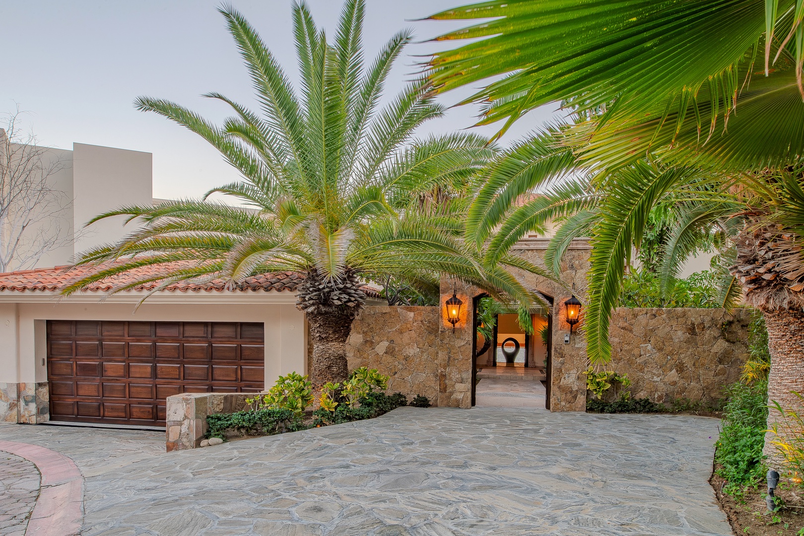 A striking stone-and-stucco facade framed by lush palms welcomes arrivals with warm lantern lighting and a terra-cotta-tiled roof.