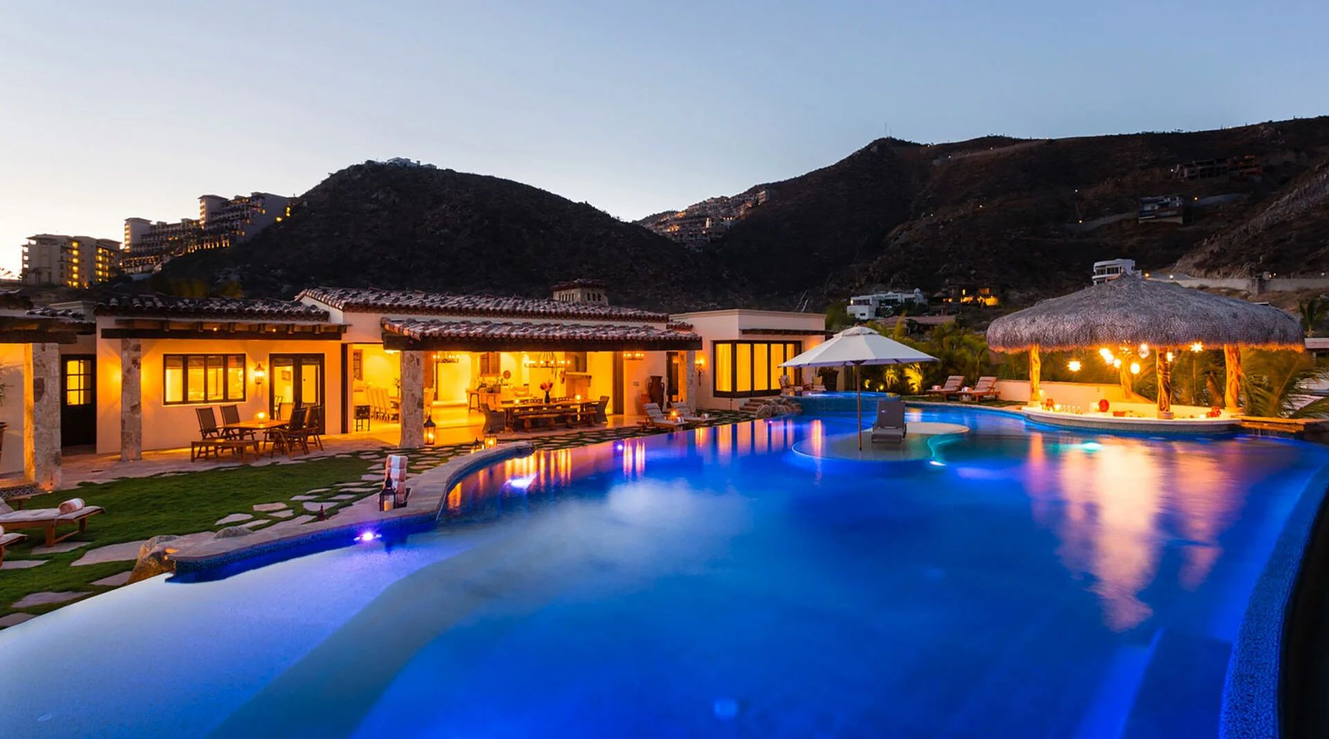 A spectacular hillside property with a stunning illuminated pool nestled between mountains. The spacious villa features covered outdoor dining areas, tropical palapas, and ambient evening lighting throughout.