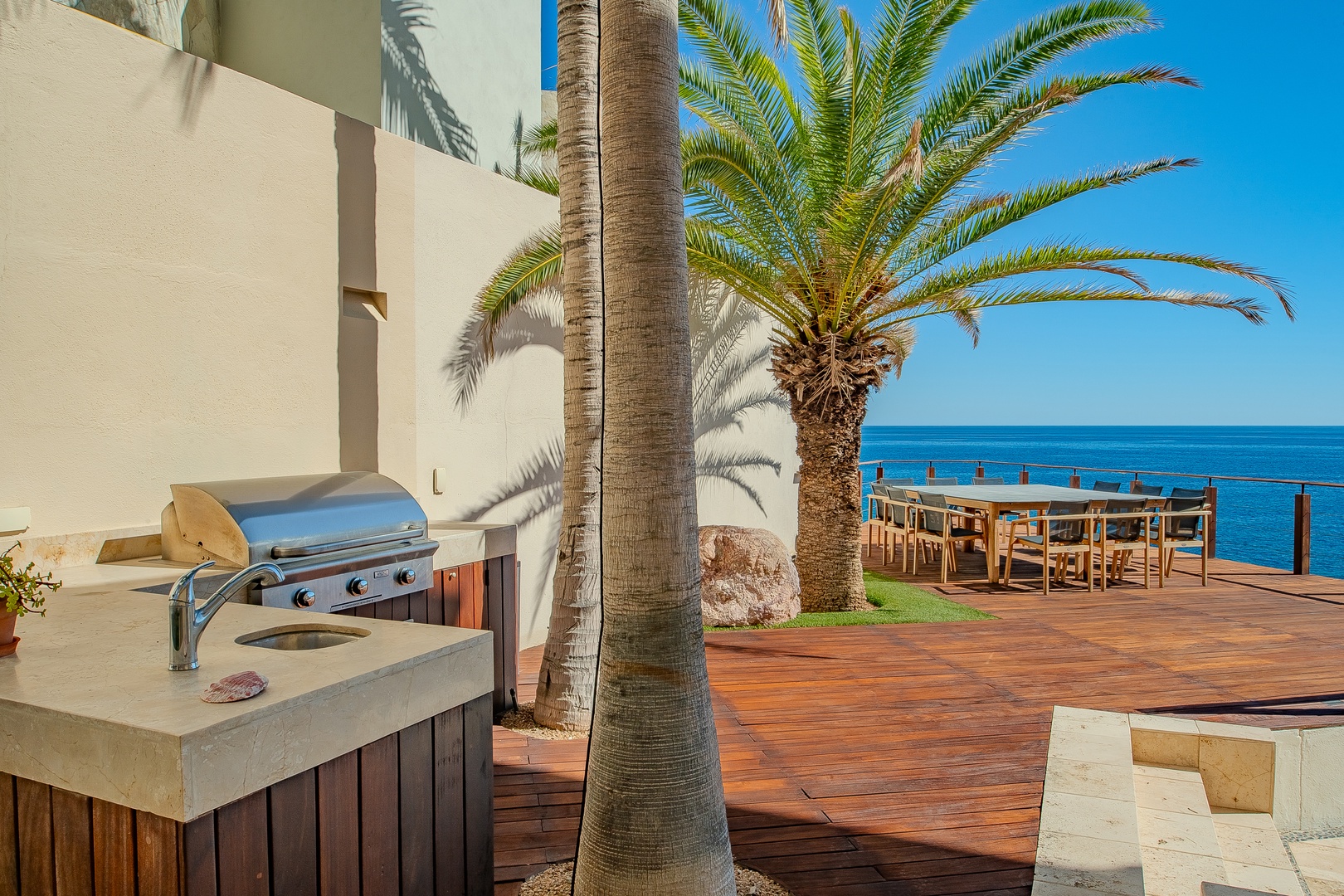 A sun-drenched oceanfront terrace with a built-in BBQ area overlooking brilliant blue waters, complete with palm trees and outdoor dining space perfect for coastal gatherings.