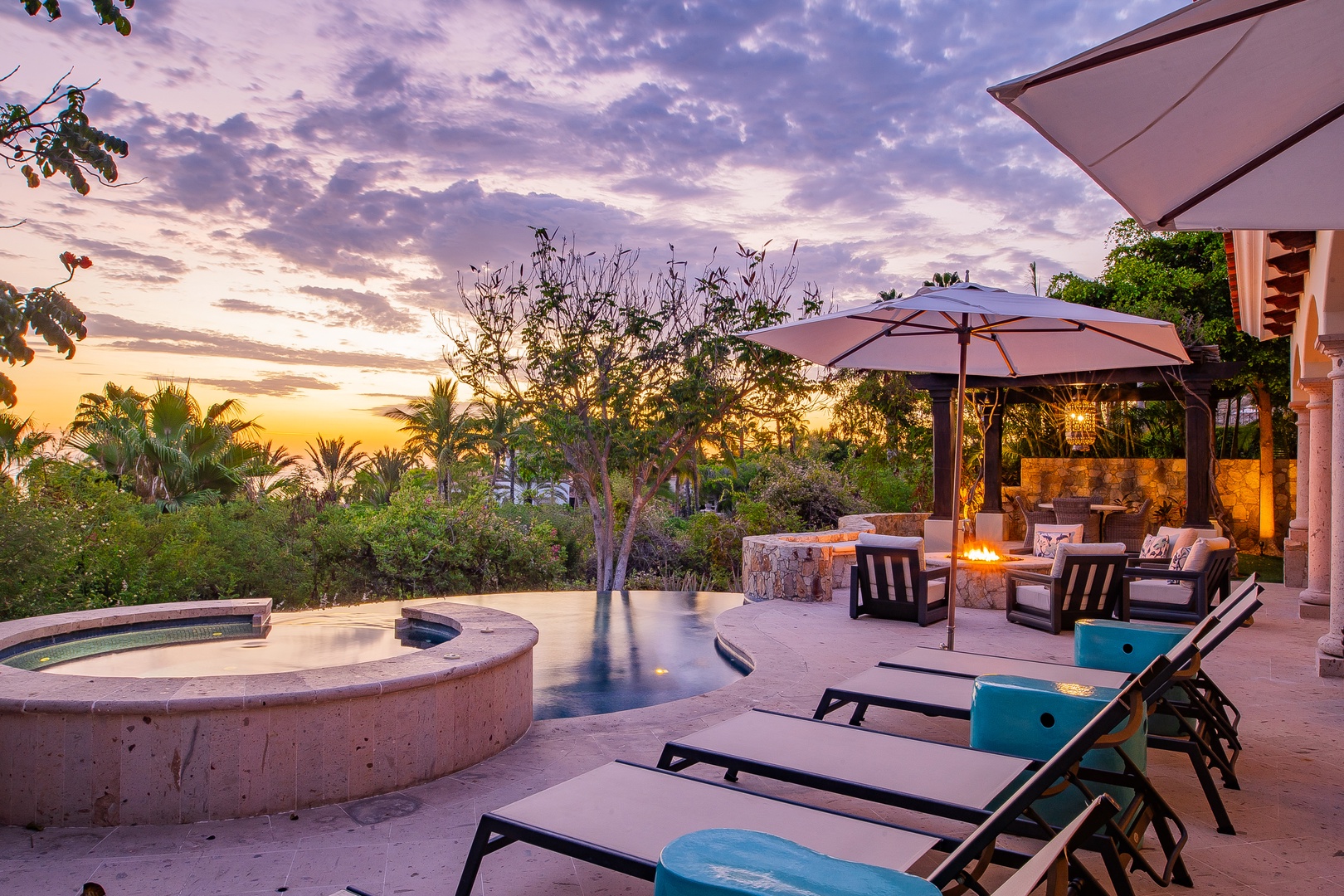 The property offers a serene outdoor setting with a heated pool, patio loungers, and lush tropical landscaping, set against stunning desert sunset views.