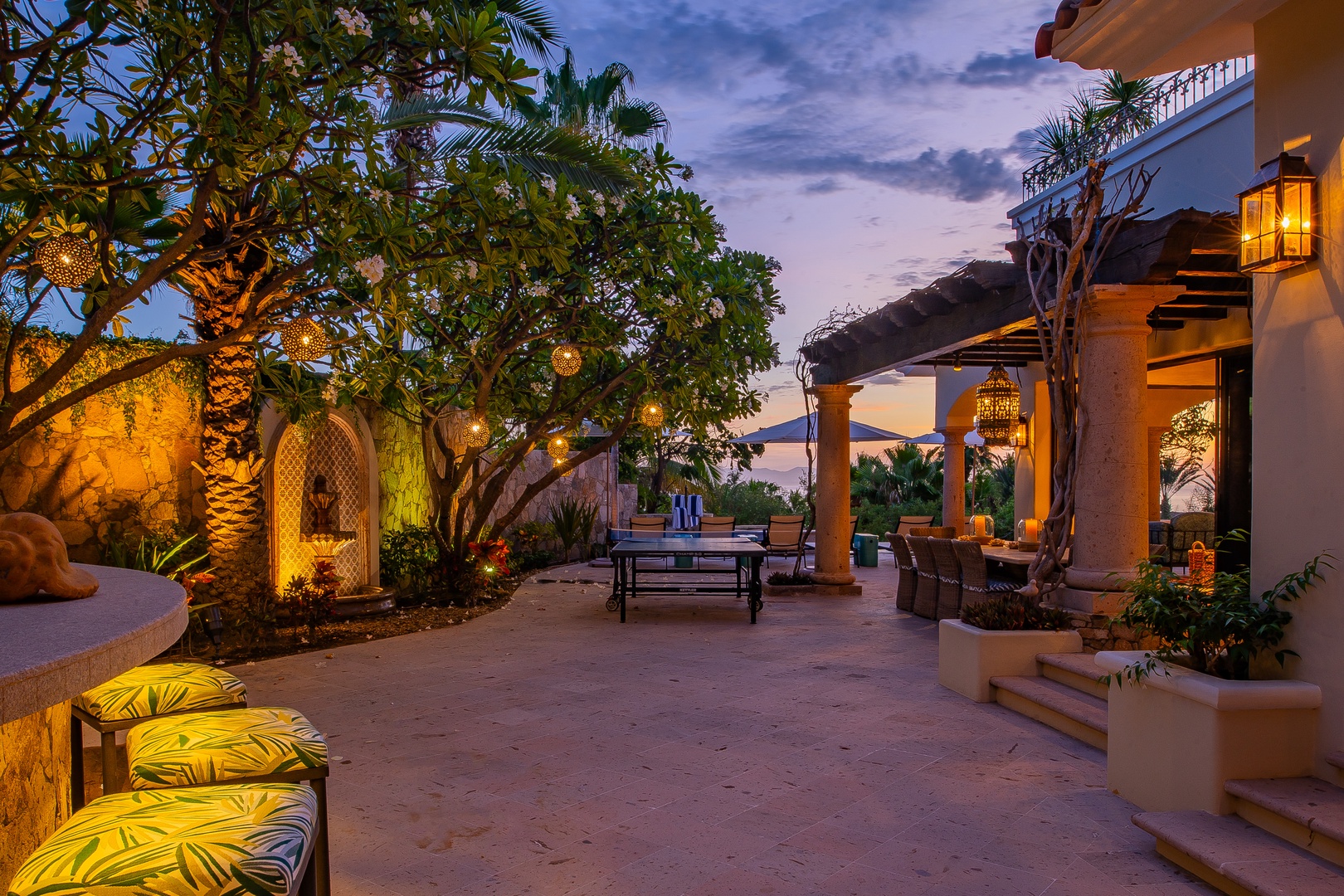 This outdoor courtyard glows as dusk settles, with lanterns casting warm light across inviting seating areas, lush palms, and serene architecture—an enchanting space for evening relaxation.