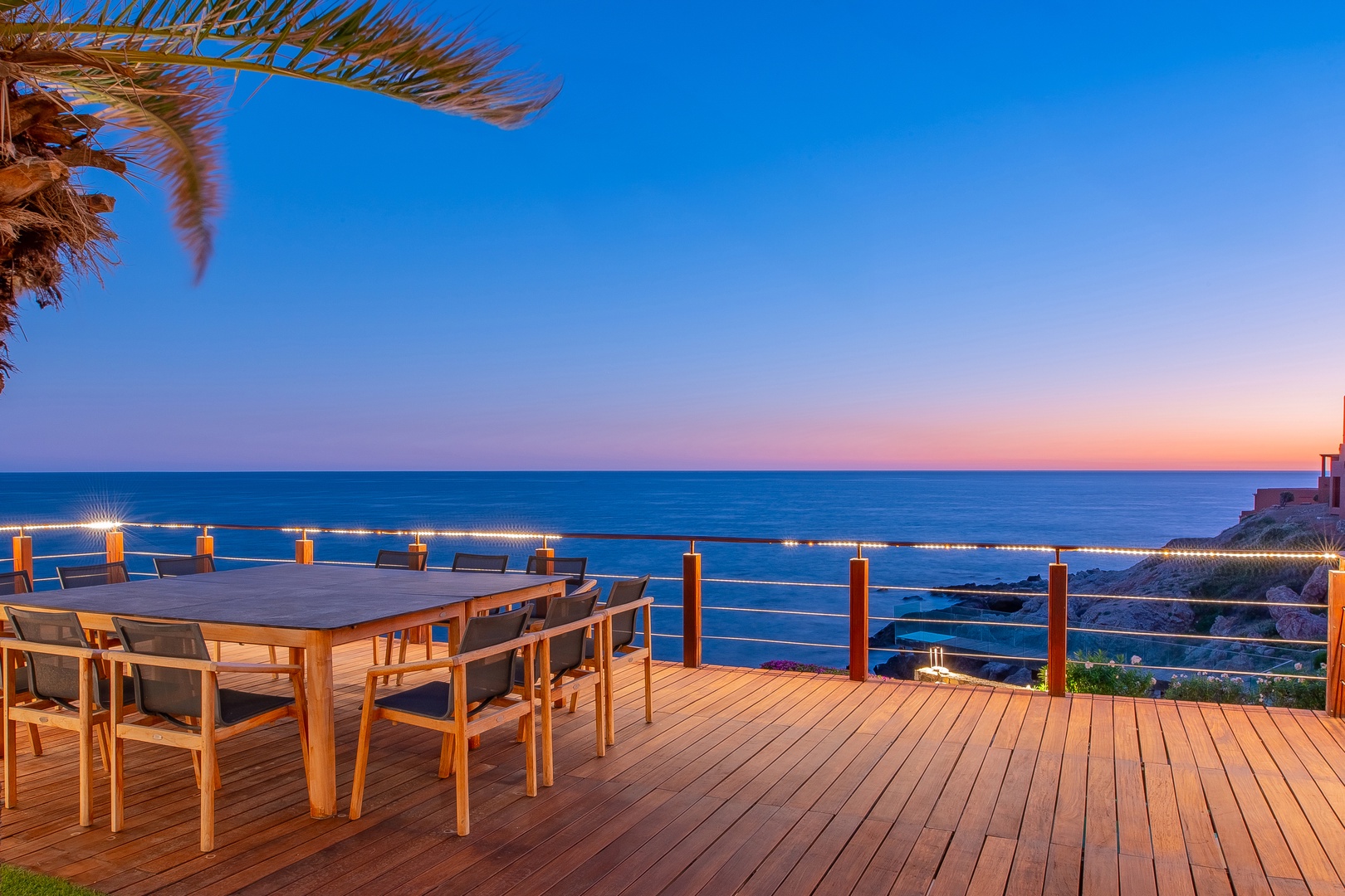 Twilight paints the sky in soft pastels beyond this oceanfront terrace—a stunning backdrop for the property. The deck, table, and string lights frame the coastal setting guests arrive to.