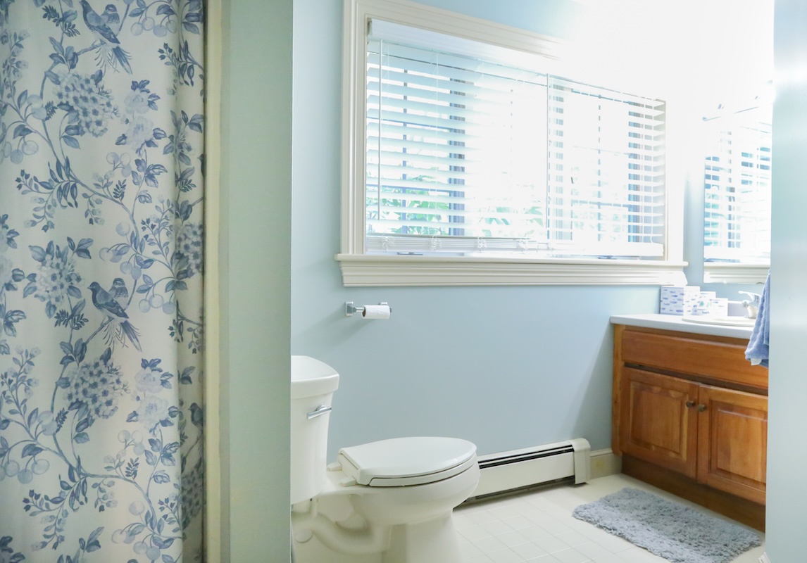 Your private bathroom offers serene coastal vibes with soft blue walls, natural light streaming through plantation shutters, and classic wood cabinetry—a calm, refreshing retreat after your day.