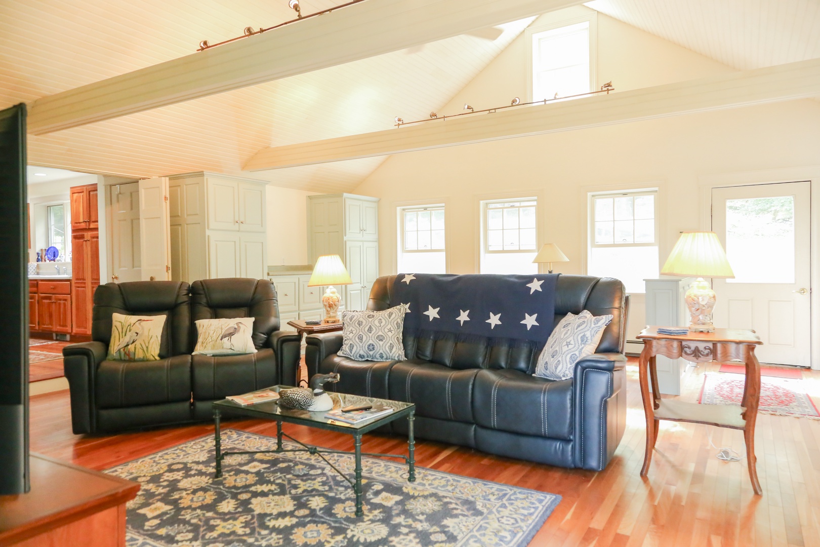 Unwind in your spacious living room with soaring vaulted ceilings and abundant natural light streaming through multiple windows. Sink into plush leather seating and let the warm wood floors and coastal decor help you settle into vacation mode.