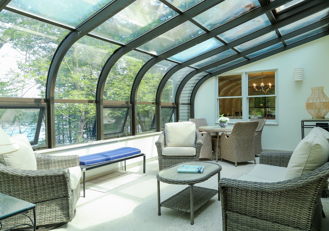 Settle into your private solarium, where curved glass walls and a skylight invite natural light to dance across comfortable wicker seating. Lush trees frame your secluded retreat, creating a peaceful sanctuary to unwind with a book or morning coffee.