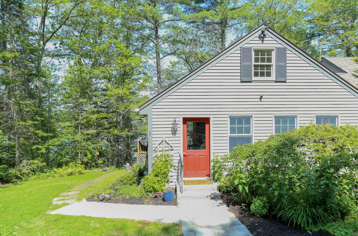 A charming cottage-style home nestled among lush greenery, featuring a welcoming coral-red front door and peaceful wooded surroundings that create a serene retreat atmosphere.