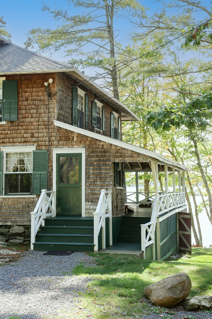 Charming cedar-shingled cottage with welcoming green trim and wraparound porch surrounded by trees.