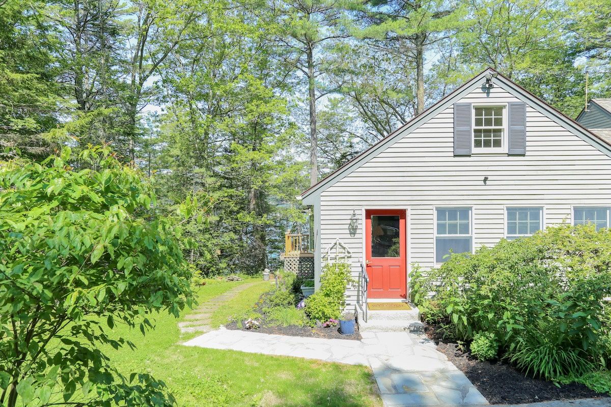 Charming cottage-style retreat nestled among tall trees, featuring a welcoming coral-red front door and surrounded by lush summer greenery and thoughtfully landscaped gardens.