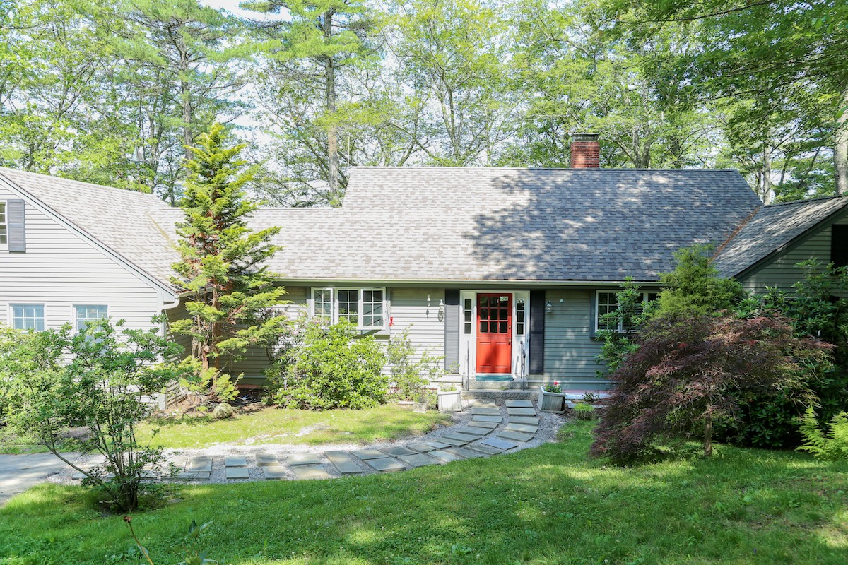 A charming cottage nestled among mature trees, featuring a welcoming red front door and stone pathway leading through a lush, landscaped yard.