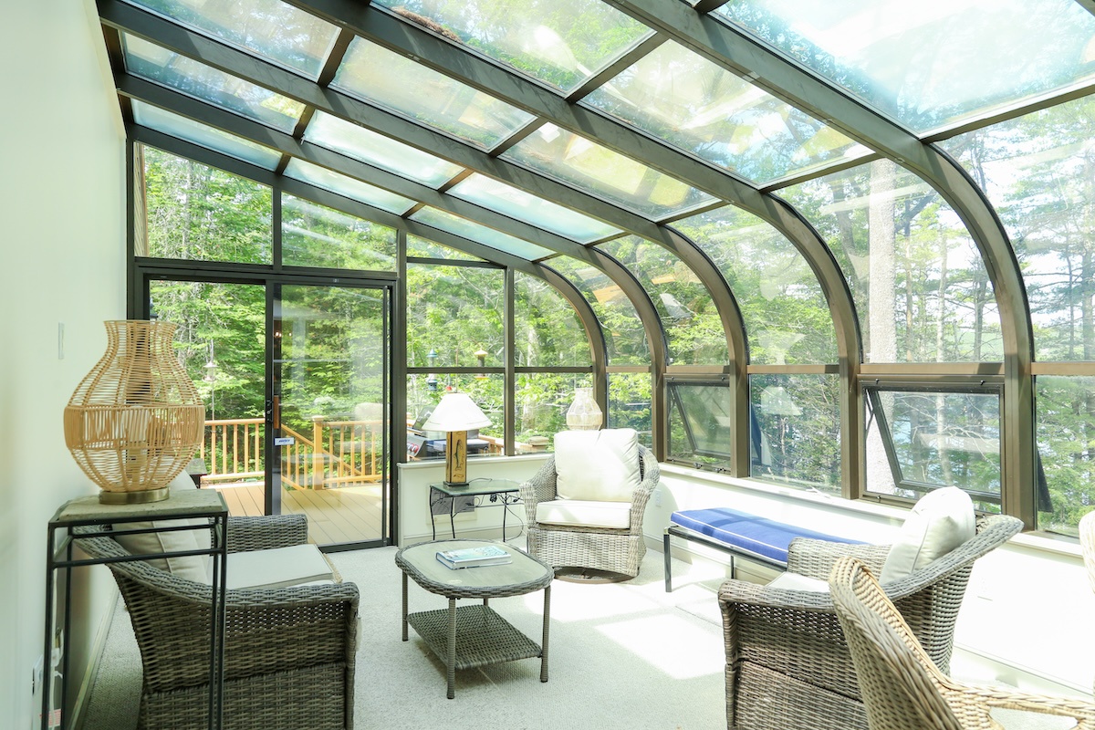 Settle into this sunlit solarium where towering glass walls and a curved ceiling frame the lush forest beyond. Sink into wicker seating as natural light floods the space, creating your peaceful retreat overlooking the private deck.