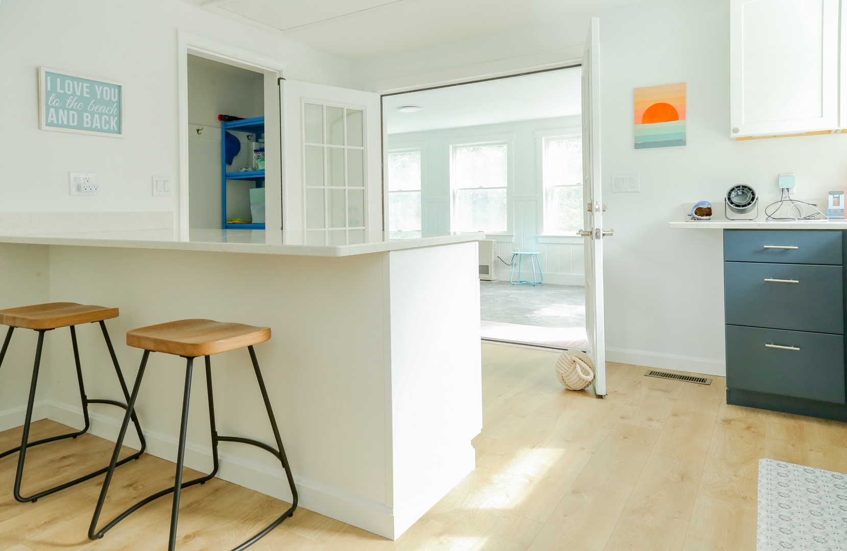 Sunlight pours through glass doors into your open kitchen, where coastal-inspired decor and modern wood accents create a breezy, beach-house vibe perfect for starting each day.
