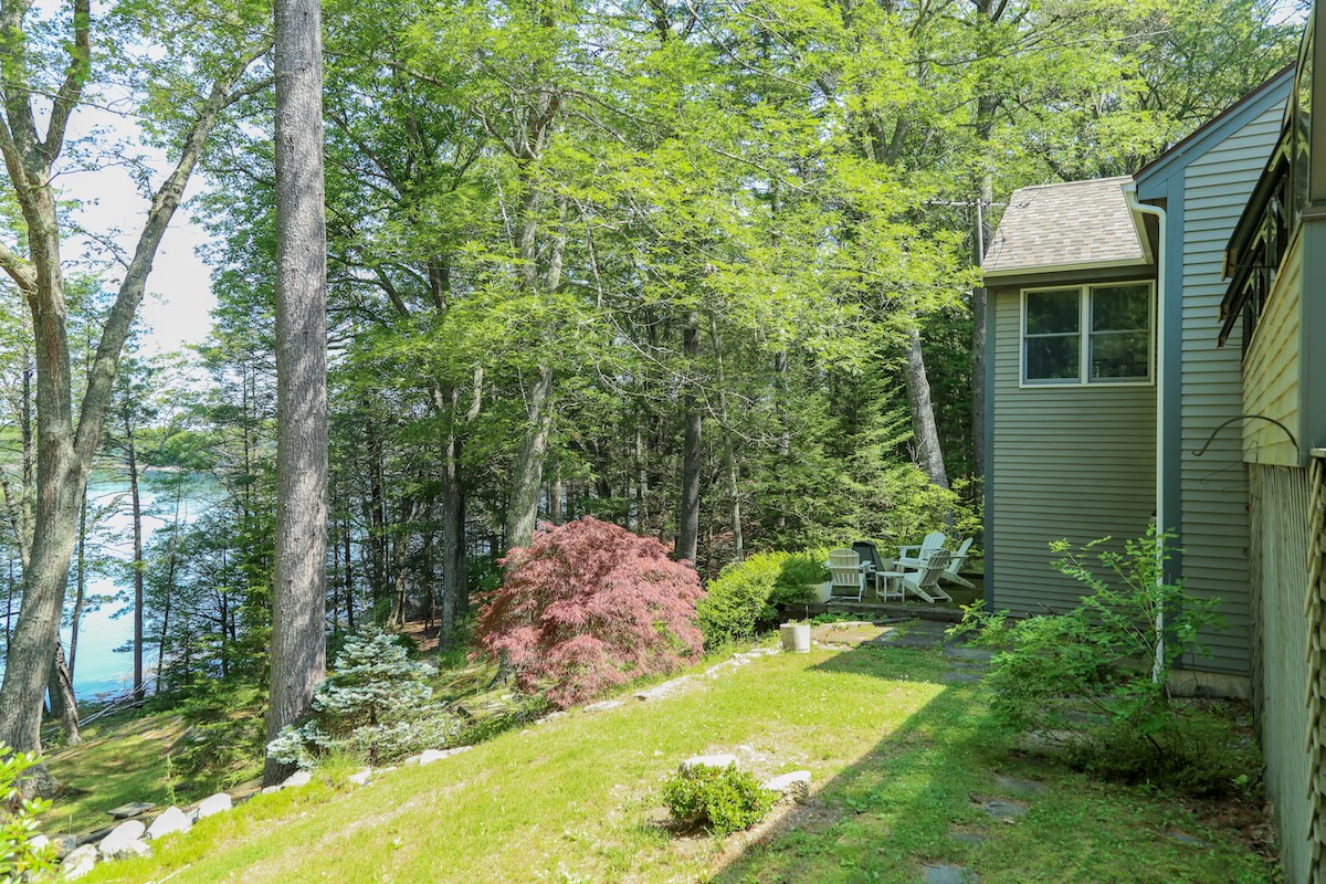 This property is nestled in a peaceful lakeside setting, surrounded by lush forest and mature trees. A charming outdoor seating area invites guests to relax amidst the natural beauty and tranquility of the wooded landscape.