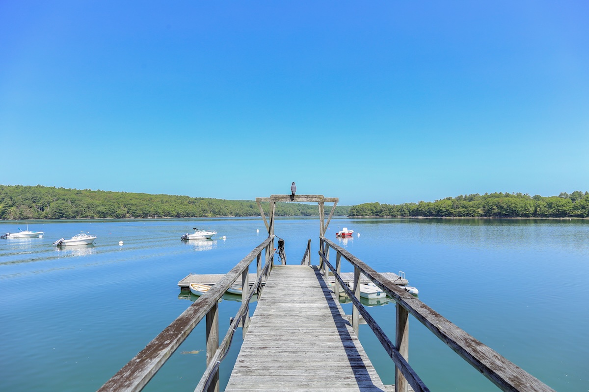 A charming wooden dock stretches over Damariscotta River, framed by lush green shorelines and peaceful boats bobbing in the distance—an idyllic coastal setting.