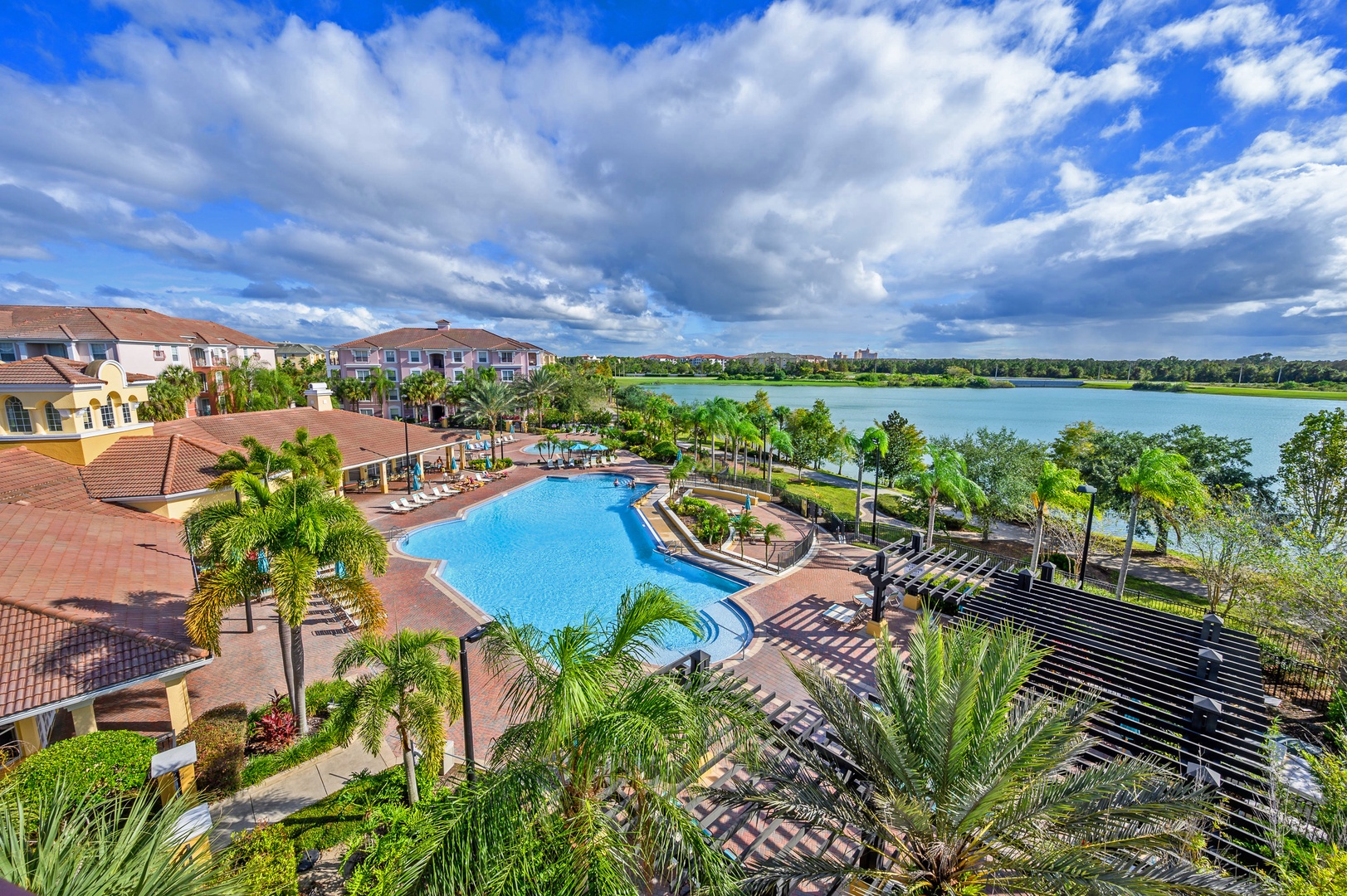 Aerial view of a lakefront resort featuring an expansive swimming pool area surrounded by lush palm trees and tropical landscaping.