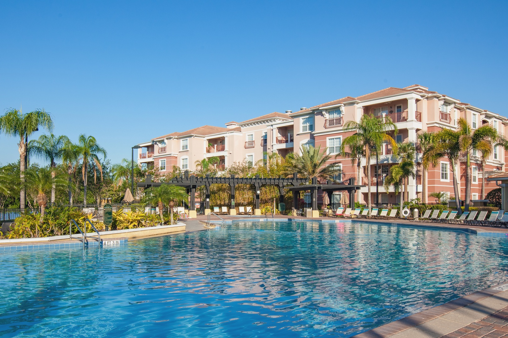 Beautiful resort-style swimming pool surrounded by tropical landscaping and Mediterranean-style buildings under clear blue skies.