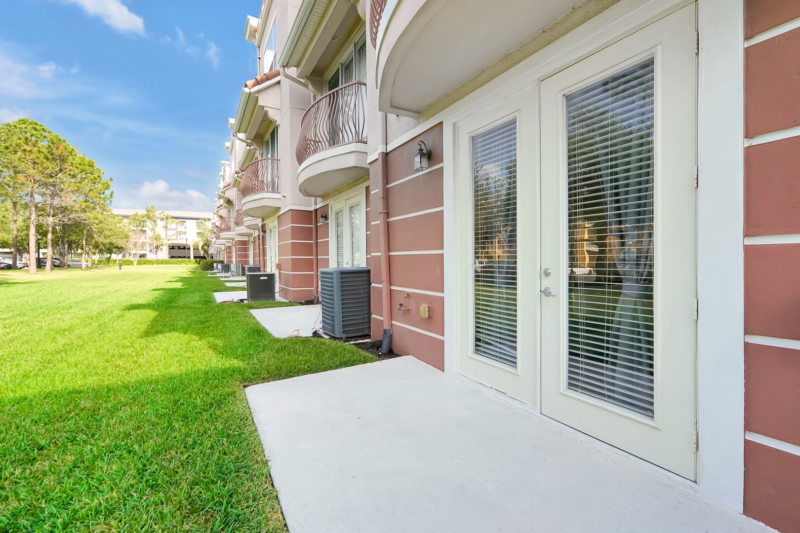 Ground-floor apartment entrance with private patio space overlooking lush lawn and landscaped community grounds under bright blue skies.