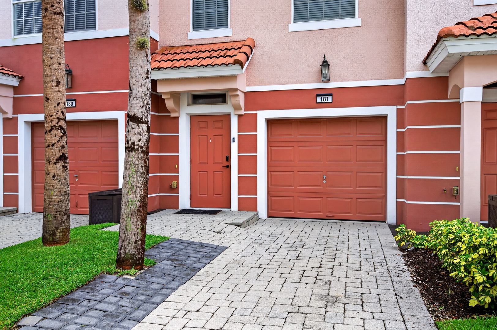 This Mediterranean-style townhome features a two-car garage and paved driveway, with terra cotta tile roofing and warm coral-toned exterior. A direct entry door leads from the garage into your unit.