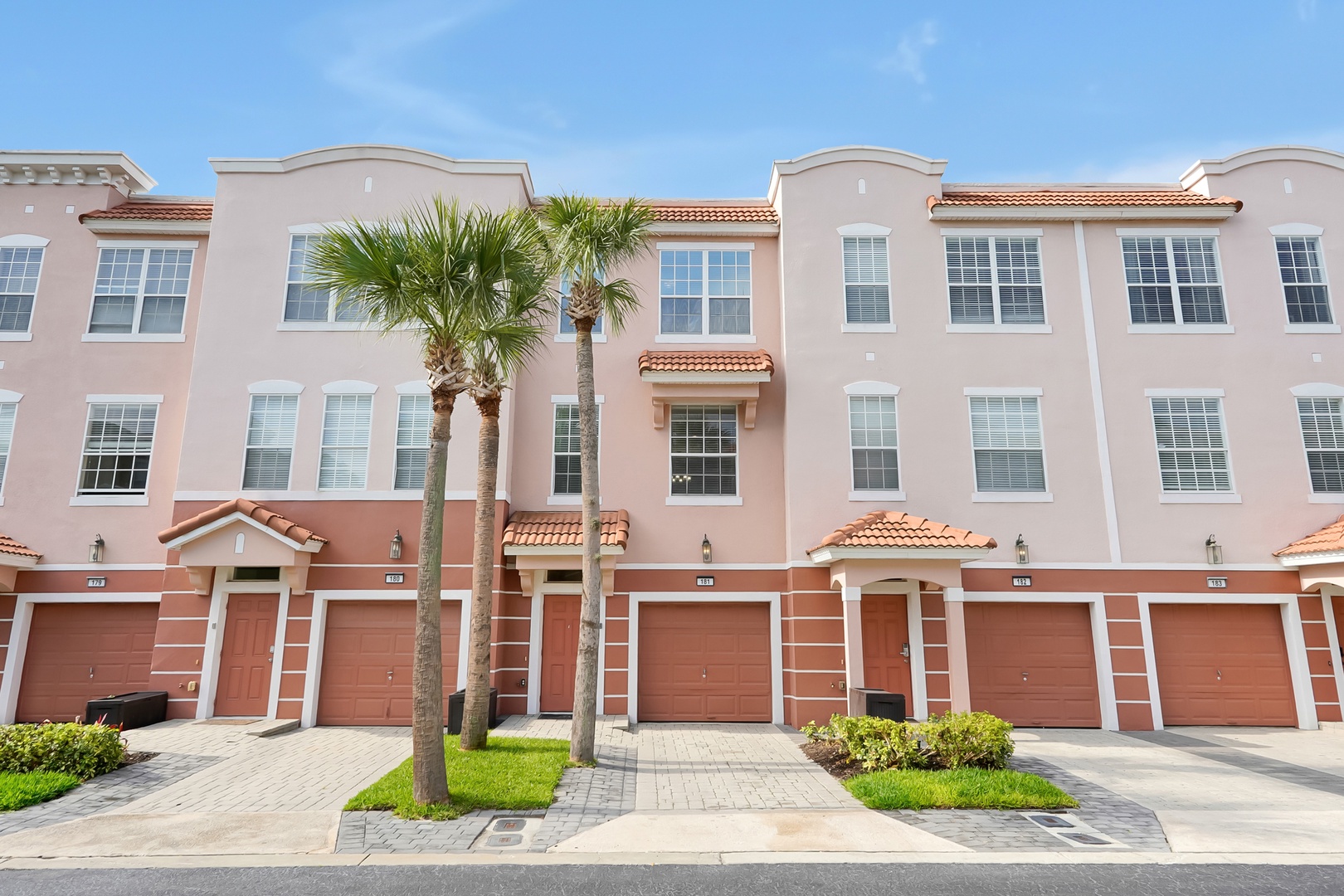 Modern townhome complex with Mediterranean-style architecture, featuring red tile roofs and palm-lined driveways in a sunny Florida setting.