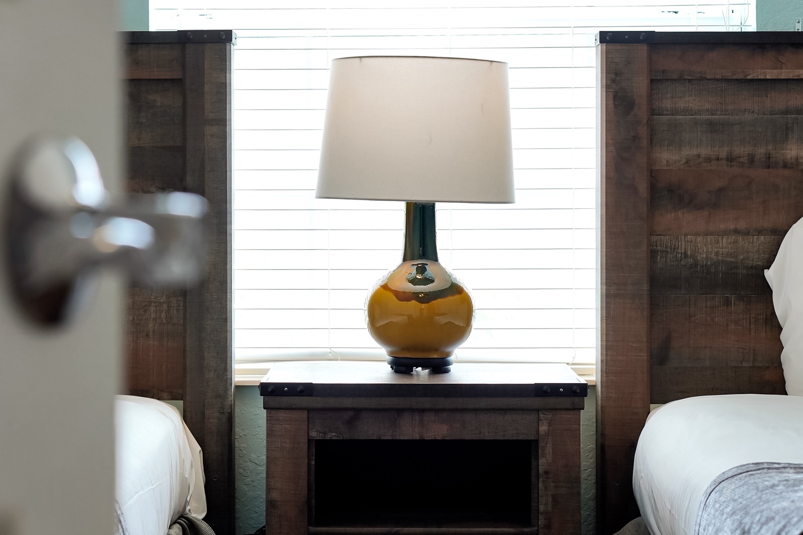 Sink into the comfort of rustic wooden beds flanking a charming nightstand, where a warm amber lamp casts a gentle glow. Fresh coastal breezes drift through the window, inviting relaxation.