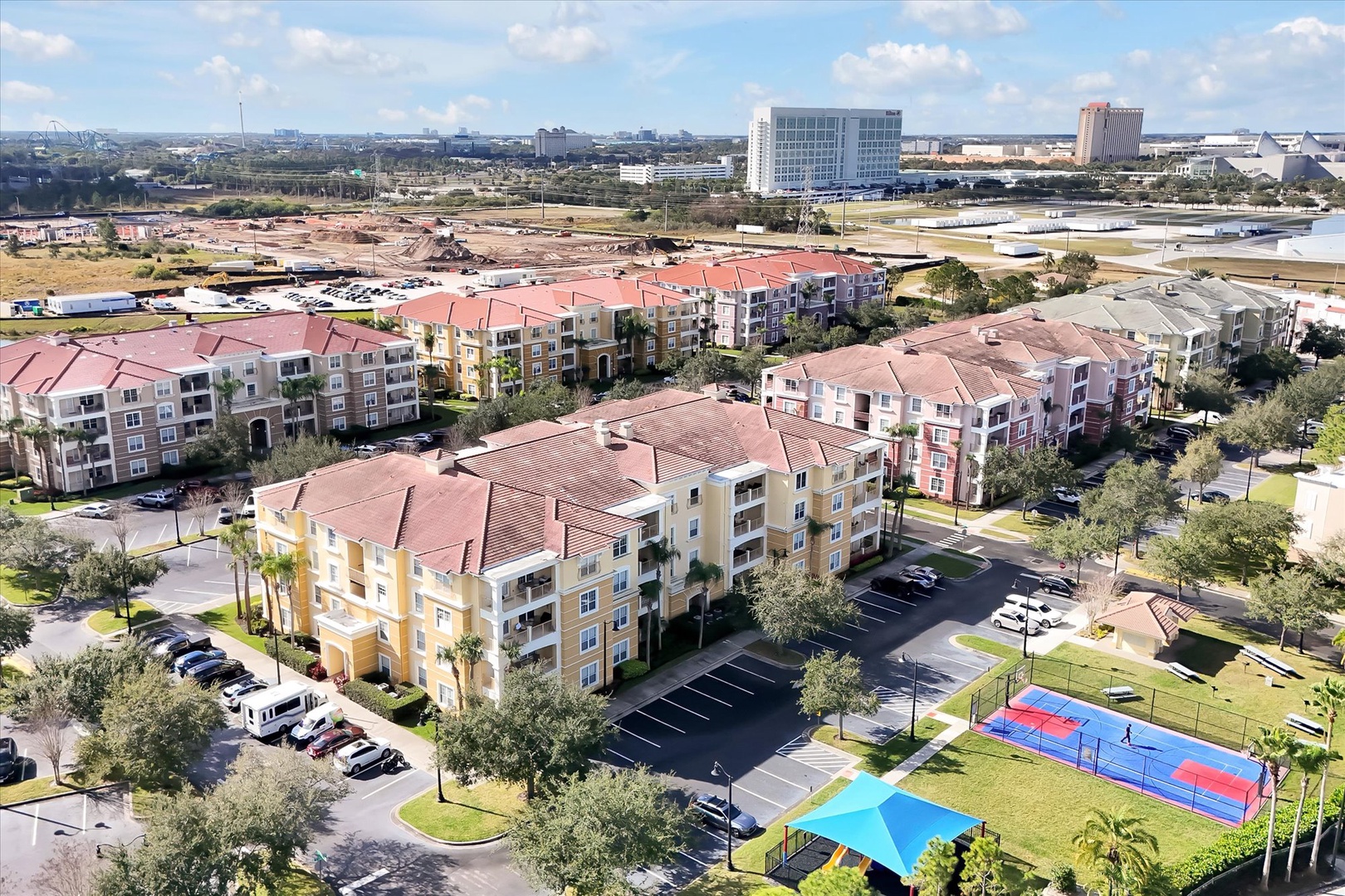 Aerial view of a residential resort community featuring colorful apartment buildings with red tile roofs, swimming pool, and tennis court.