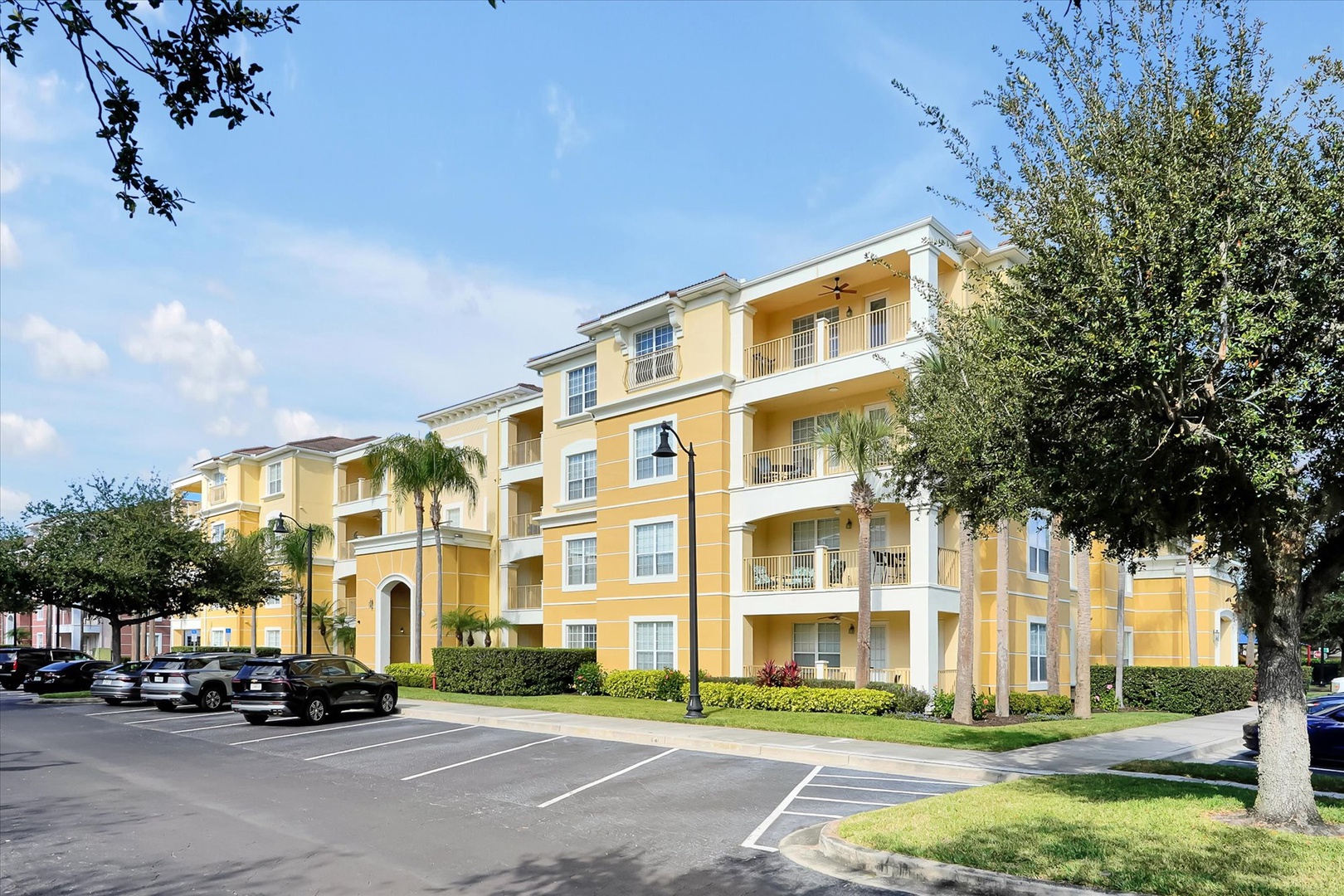 Bright yellow multi-story buildings with white trim create a cheerful Mediterranean-style complex surrounded by lush tropical landscaping and convenient parking.