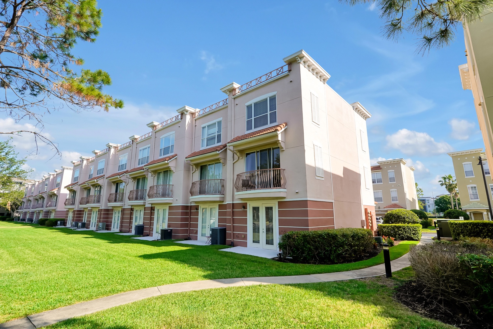 Modern townhouse complex with manicured gardens and sunny skies creates a welcoming arrival experience in a peaceful residential setting.